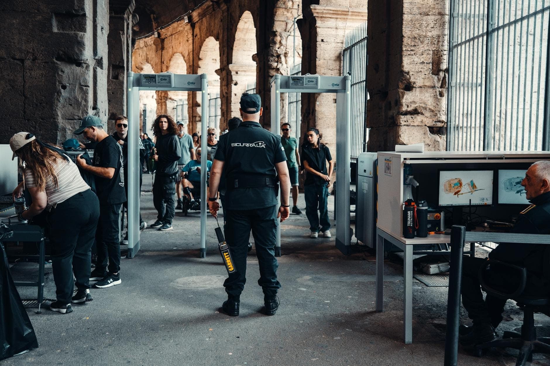 Turisti che passano attraverso i controlli di sicurezza con metal detector e macchine a raggi X all'interno di un monumento romano, sorvegliati dal personale di sicurezza.