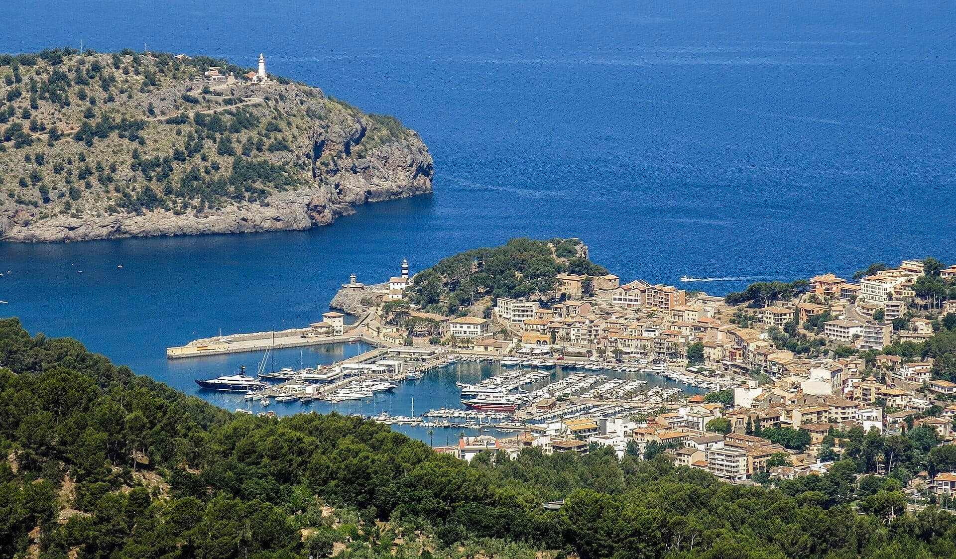 Luftaufnahme des Port de Sóller mit einem bootsgefüllten Yachthafen, mediterranem blauem Wasser, grünen Hügeln und einem historischen Leuchtturm.