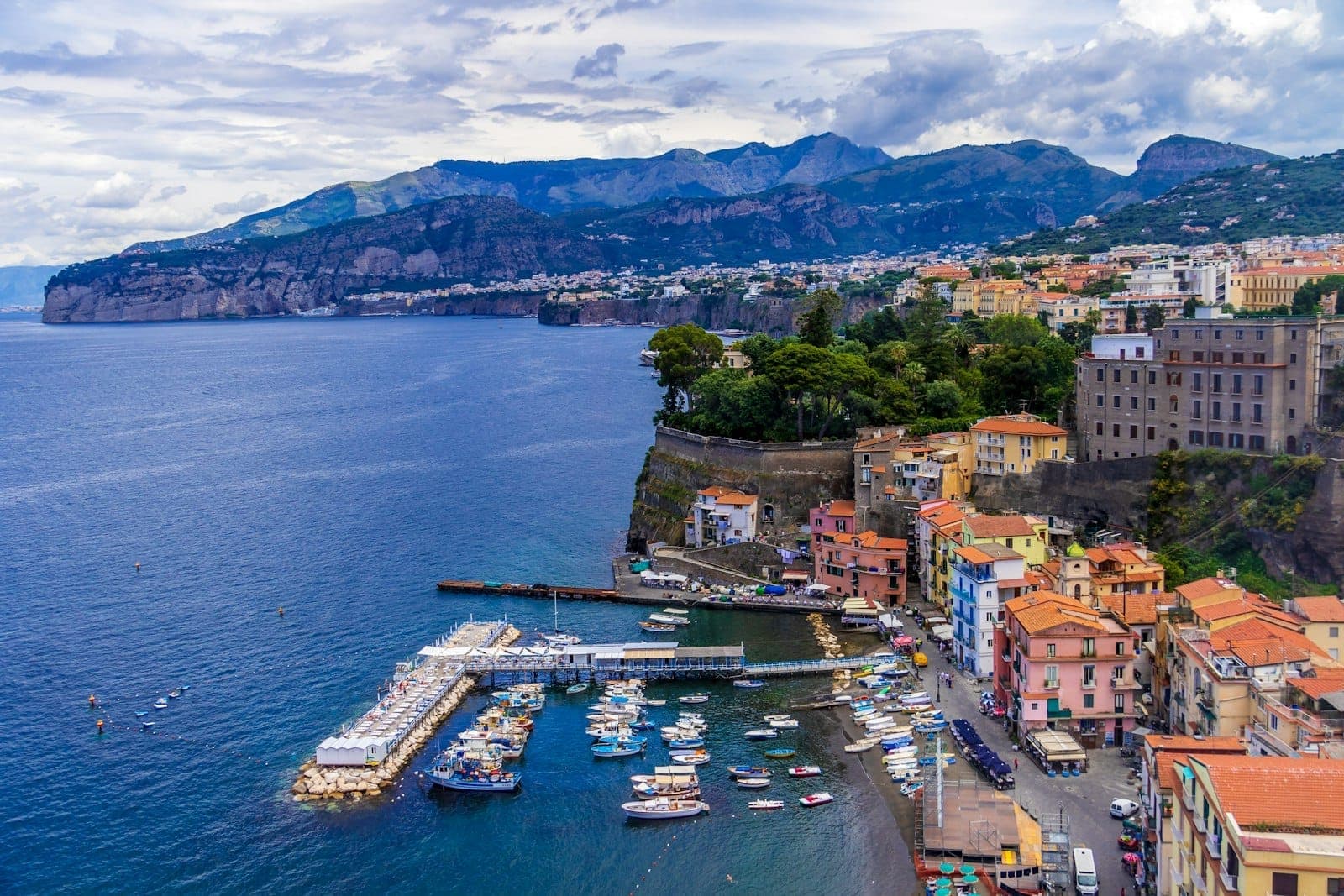 Vista aérea dos coloridos edifícios à beira-mar de Sorrento, pequenos barcos atracados na marina e penhascos escarpados sobre a Baía de Nápoles sob céus nublados.