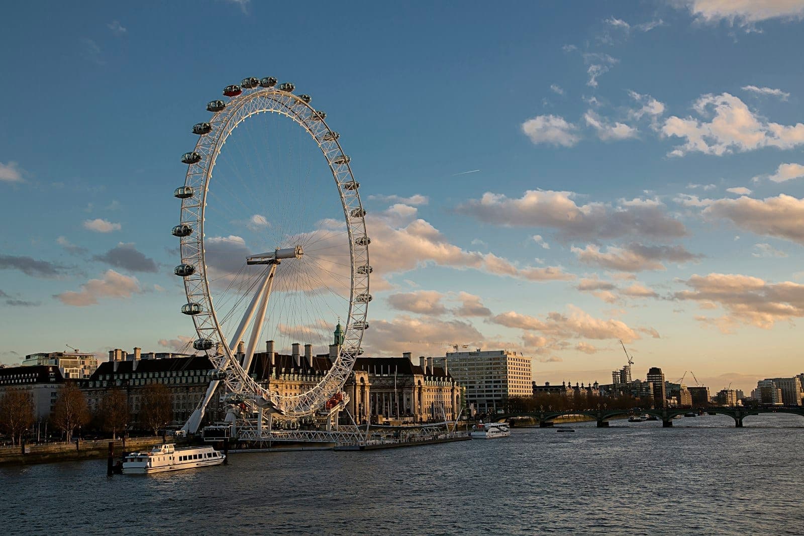 London Eye i pobliskie budynki na South Bank skąpane w ciepłym wieczornym świetle, z Tamizą na pierwszym planie i częściowo zachmurzonym błękitnym niebem.