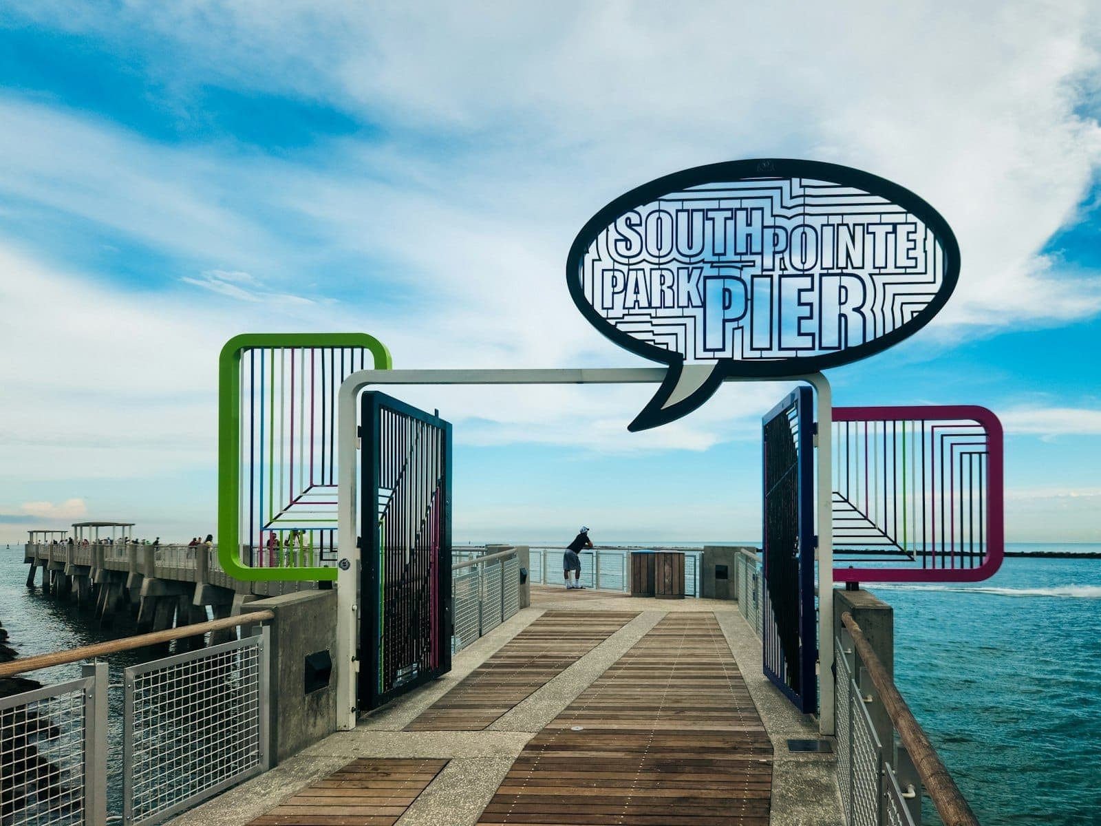Entrance to South Pointe Park Pier in Miami Beach, featuring colorful art, a large sign, clear blue sky, and calm ocean waters.