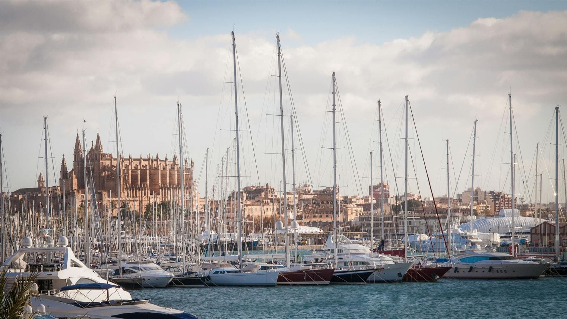 Marina mit Segelbooten im Vordergrund, dahinter die Kathedrale von Palma und die Stadtsilhouette unter einem bewölkten blauen Himmel.