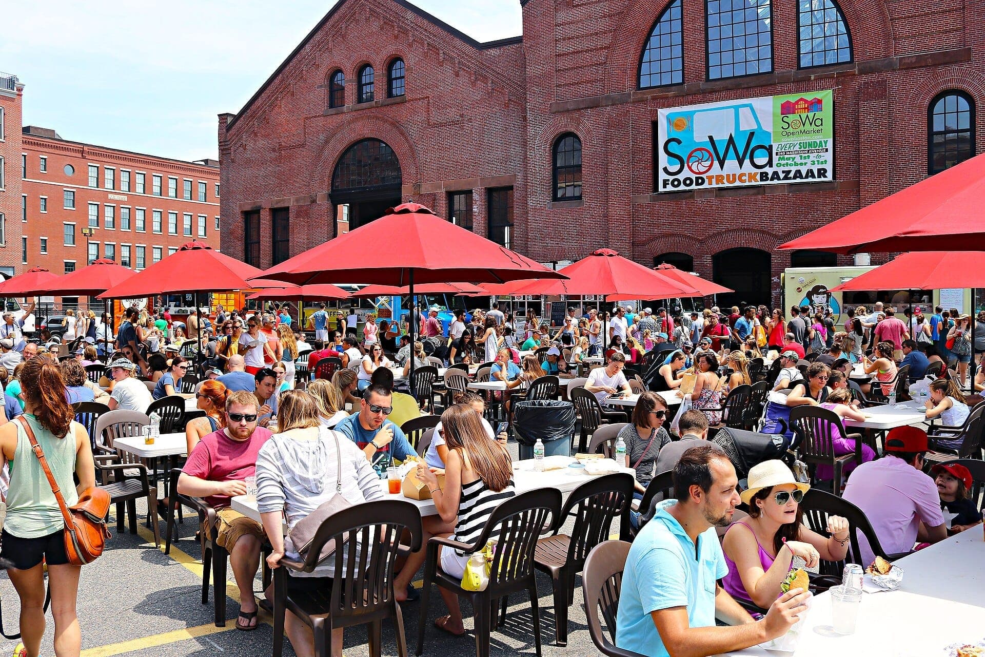 Foule profitant d'un repas en plein air sous des parasols rouges au SoWa Art + Design District, avec des bâtiments en briques et des food trucks en arrière-plan.