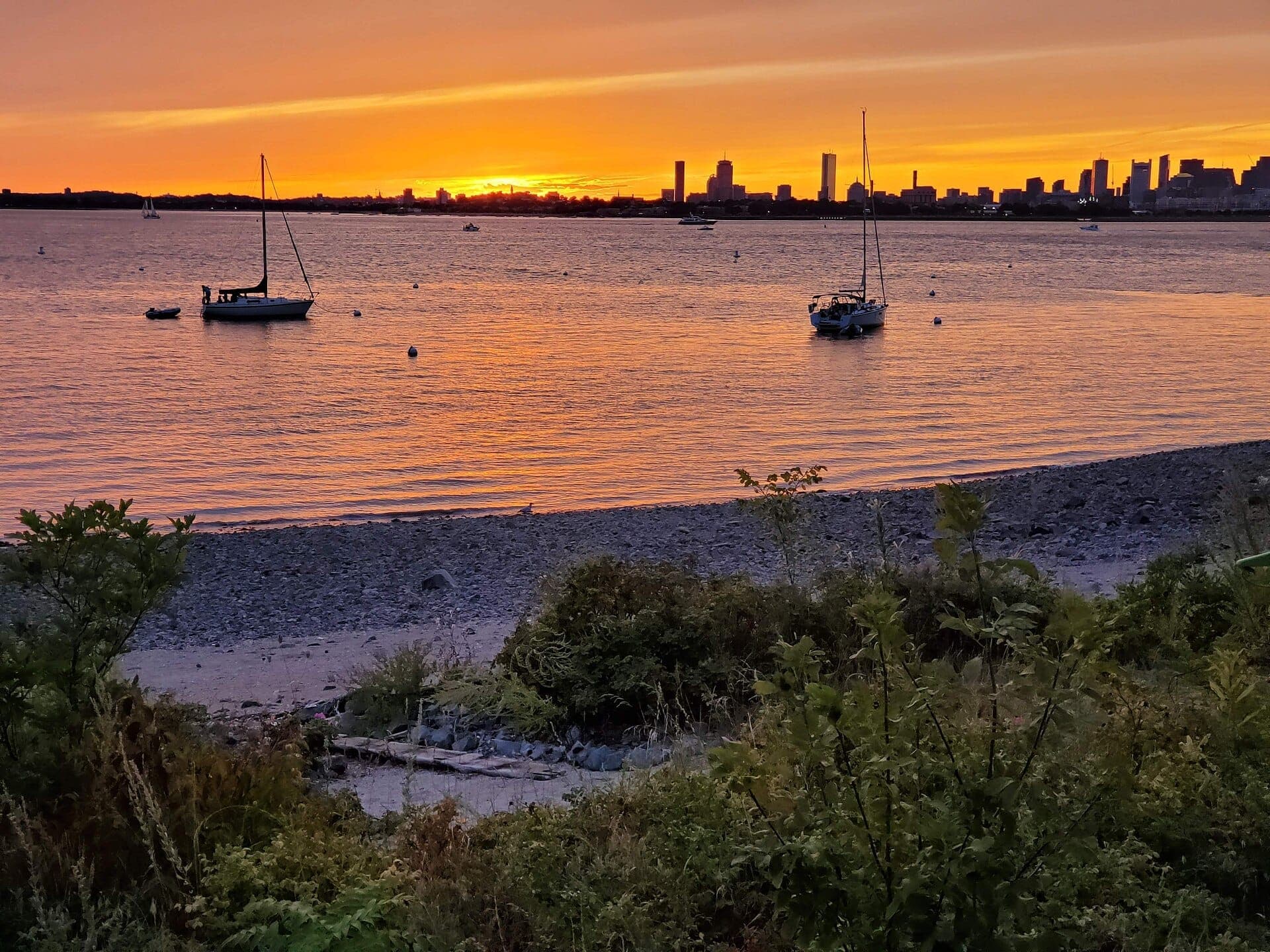 Plage de Spectacle Island avec des voiliers sur une eau calme, la skyline de Boston en arrière-plan et un coucher de soleil orange vibrant illuminant le ciel.