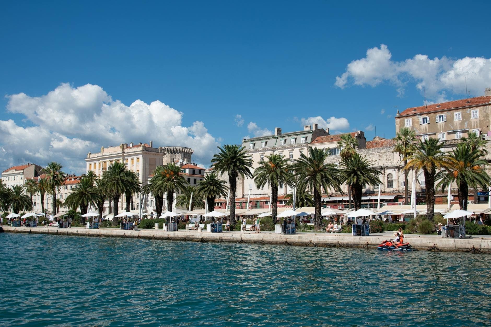 Lungomare soleggiato di Spalato fiancheggiato da palme, edifici storici e caffè all'aperto sotto un cielo azzurro con nuvole sparse.