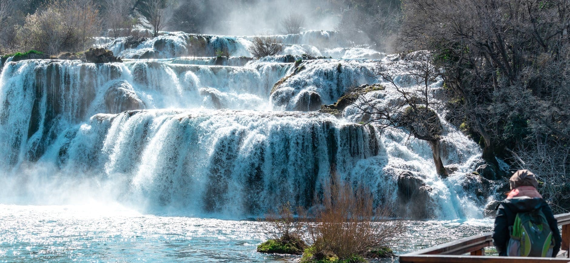 Plusieurs niveaux de cascades du Parc National de Krka dévalent des rebords rocheux avec de la brume qui monte, entourés d'arbres nus en hiver et d'eau en mouvement.