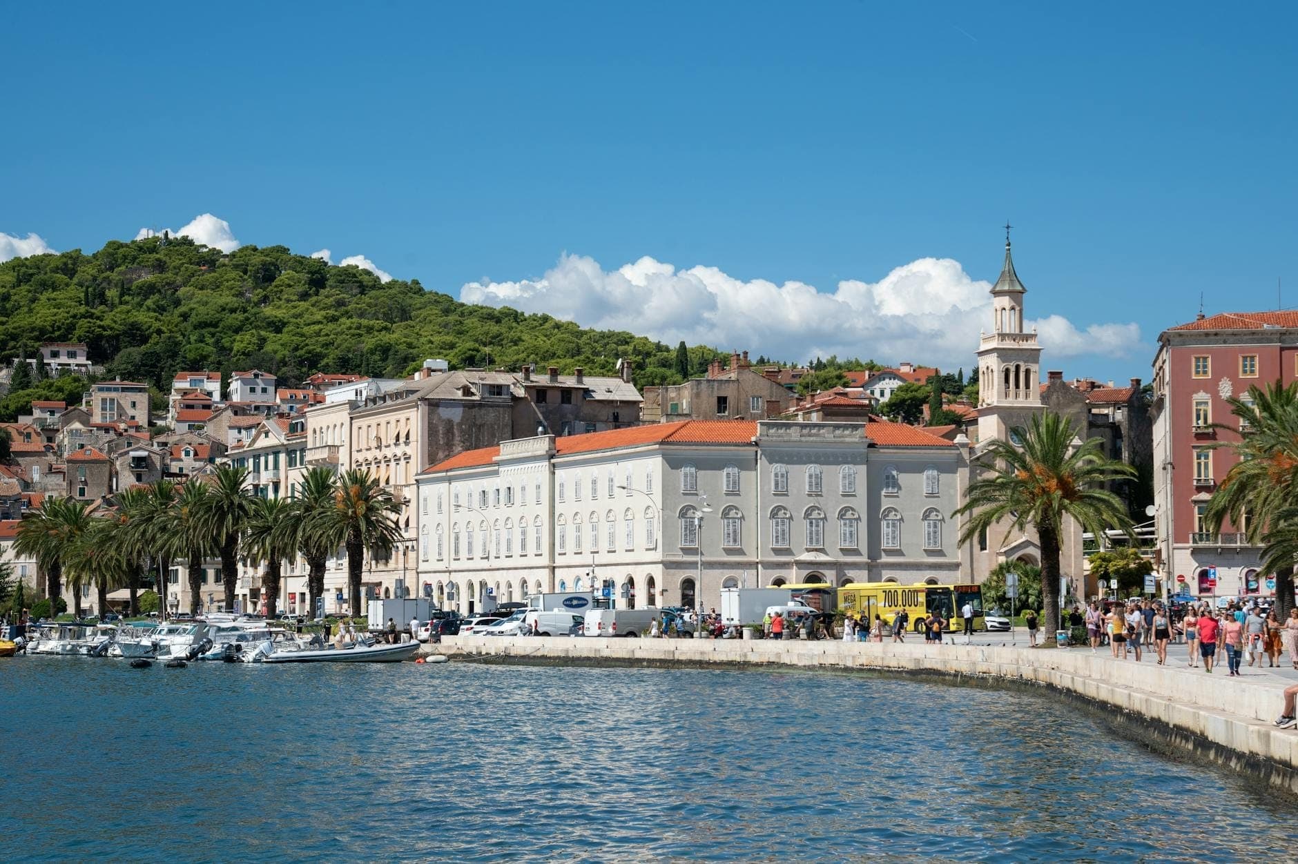 Vue de jour de la promenade Riva de Split, du front de mer, des palmiers et des bâtiments historiques avec des personnes se promenant sur le chemin piétonnier.