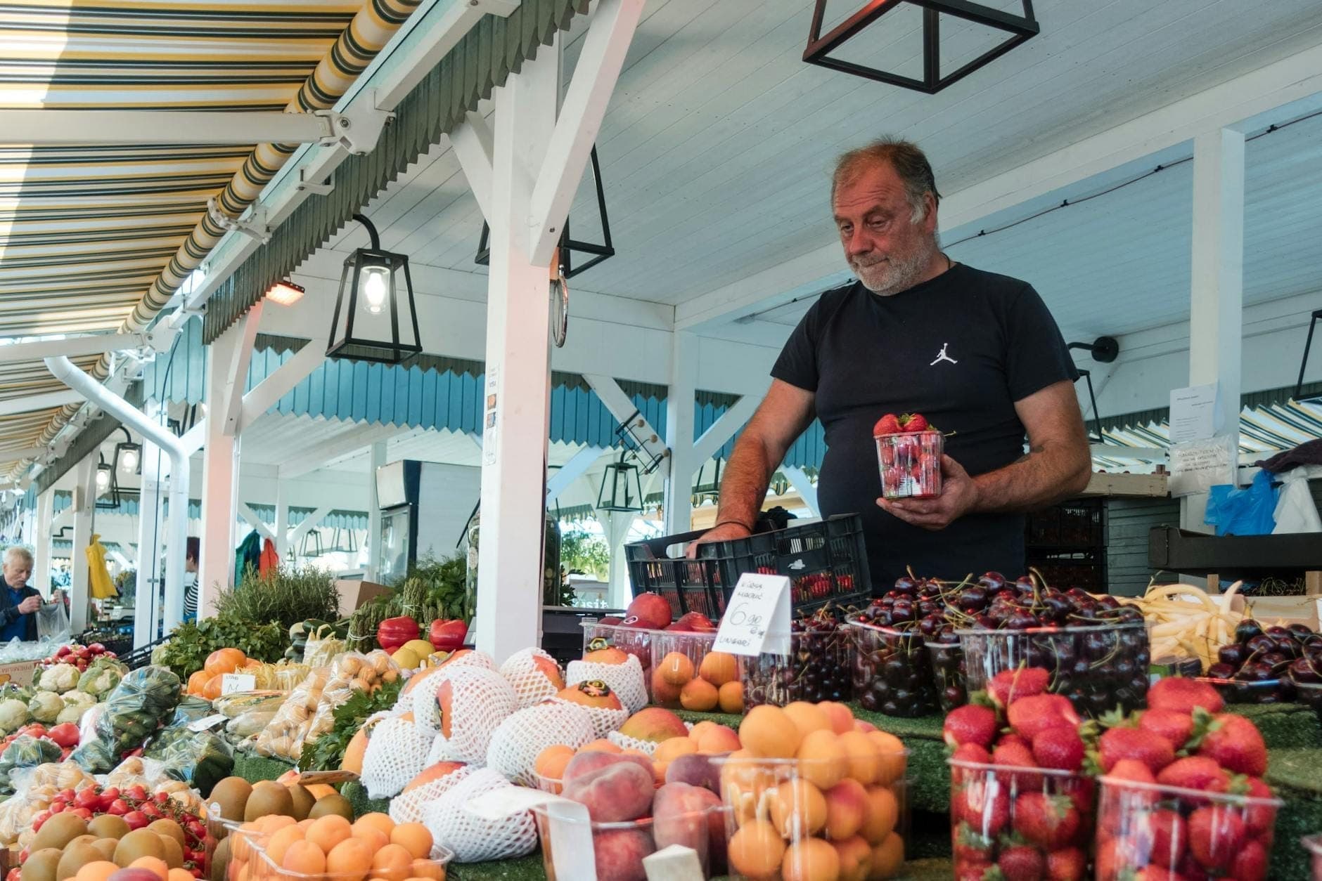 Un vendeur arrange des fruits frais sur un étal de marché en plein air sous un auvent, avec des paniers de fraises et de cerises au premier plan.
