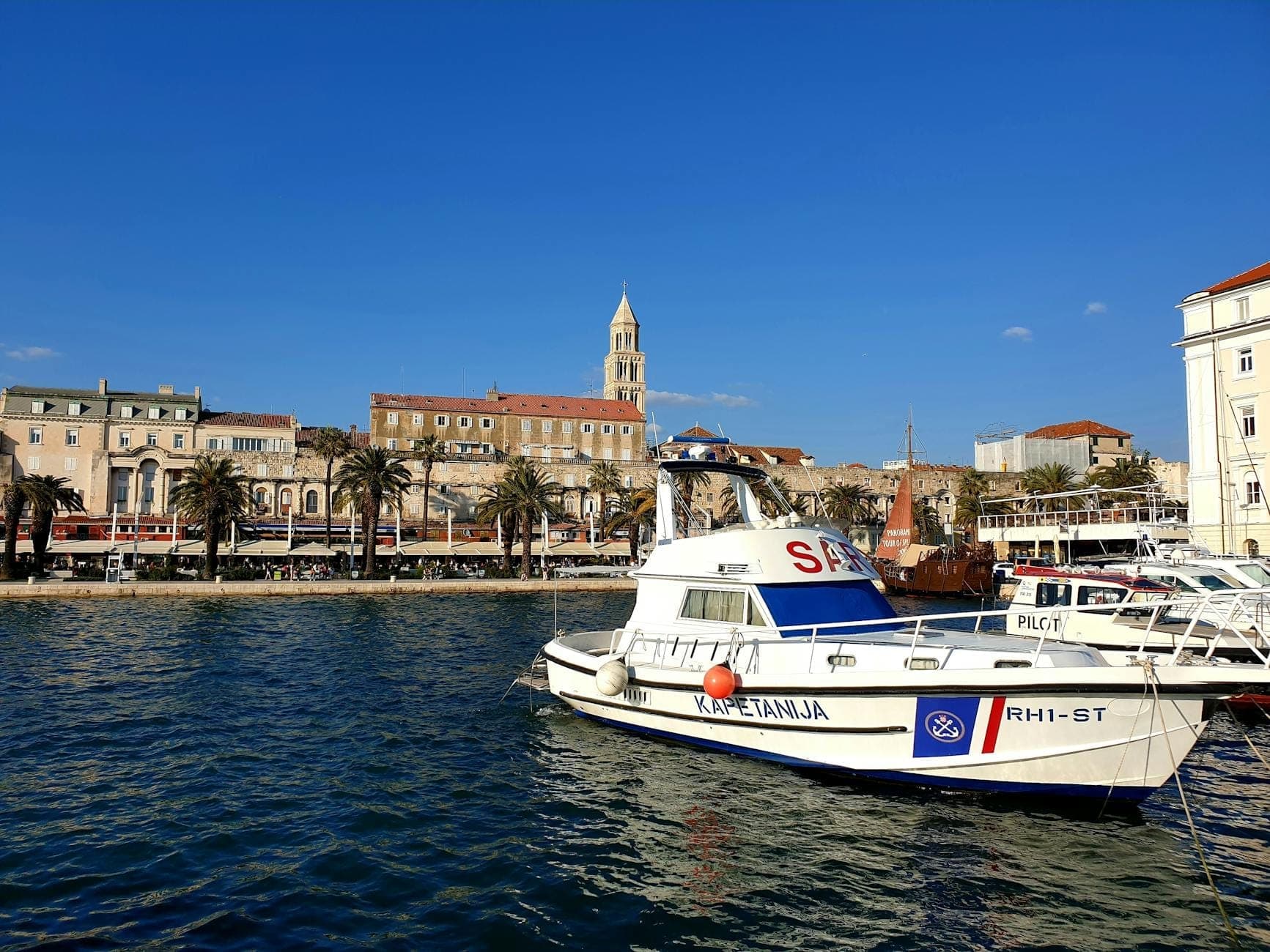Scène du port de Split avec un bateau de police ou de services d'urgence amarré devant les bâtiments historiques du front de mer et le clocher de la cathédrale.