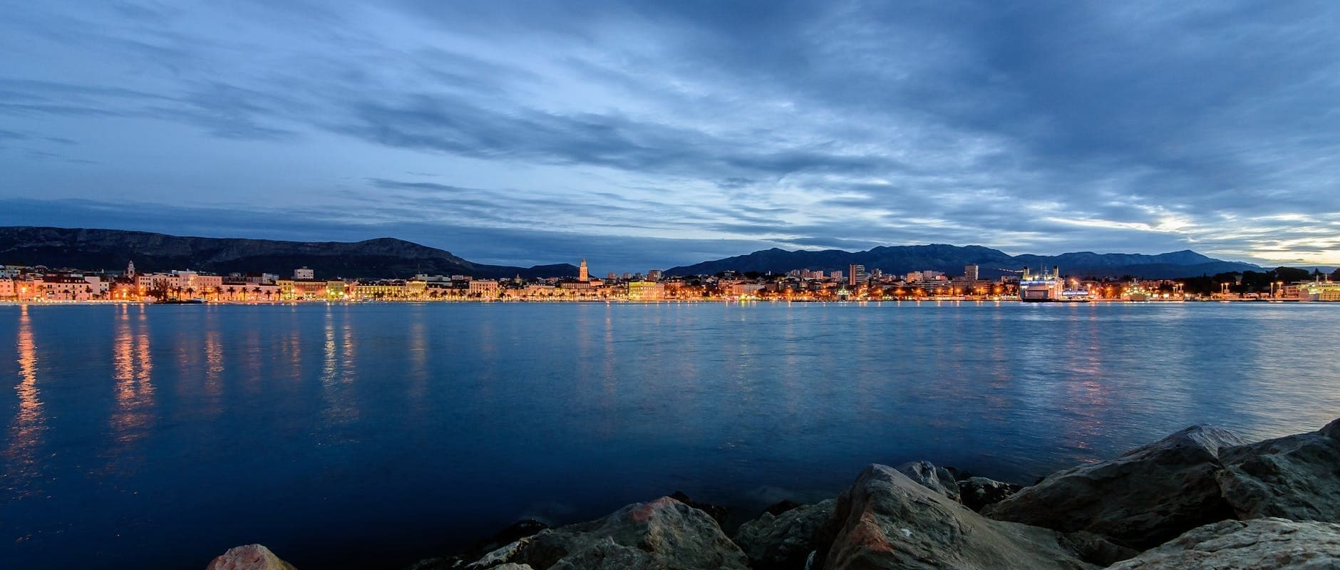 Vue panoramique en soirée du front de mer de Split avec les lumières de la ville se reflétant sur l'eau, des montagnes en arrière-plan et une ambiance calme pour la vie nocturne.