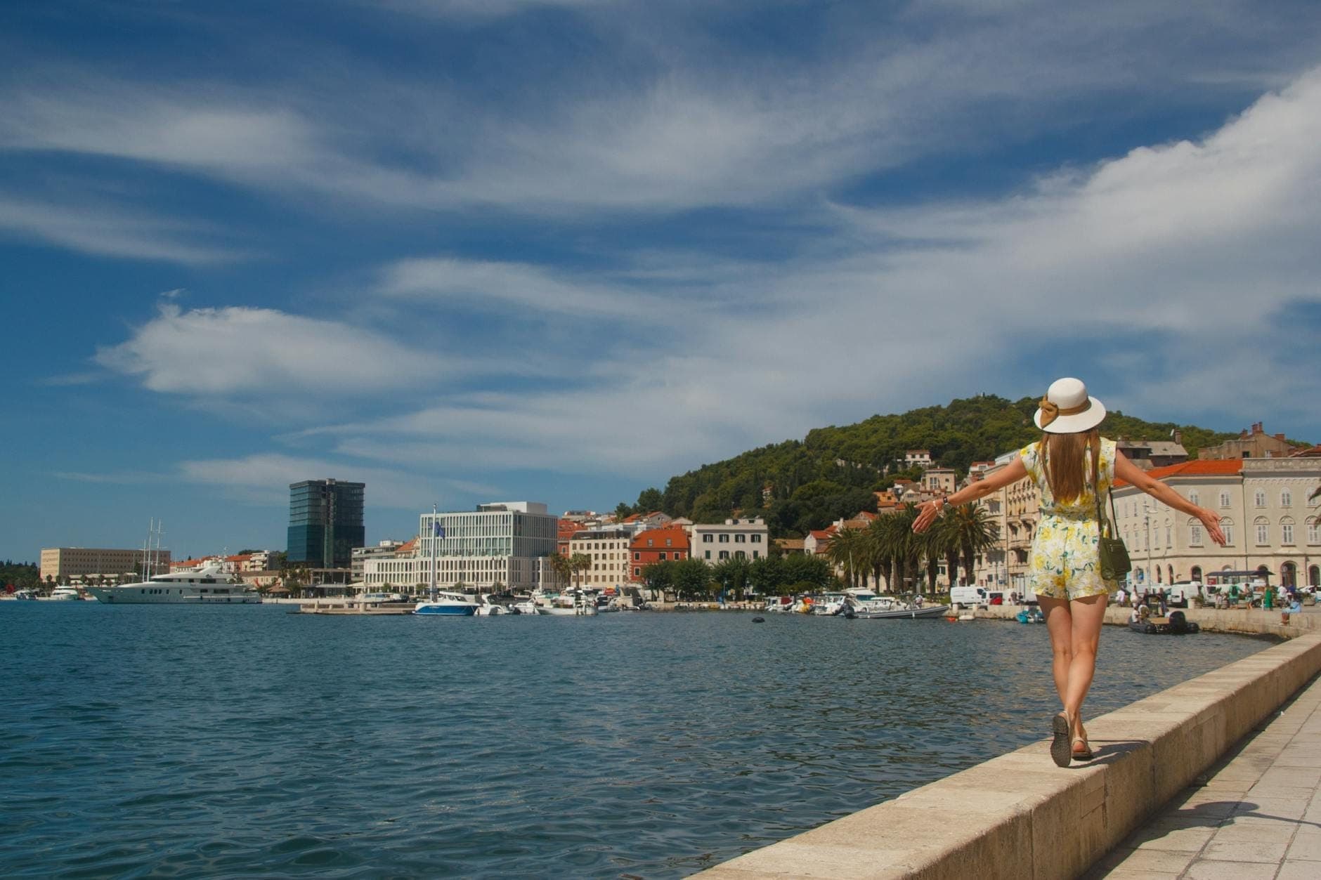 Femme se promenant seule le long de la promenade de Split au bord de l'eau, avec le paysage urbain reconnaissable et la marina en arrière-plan sous un ciel ensoleillé.