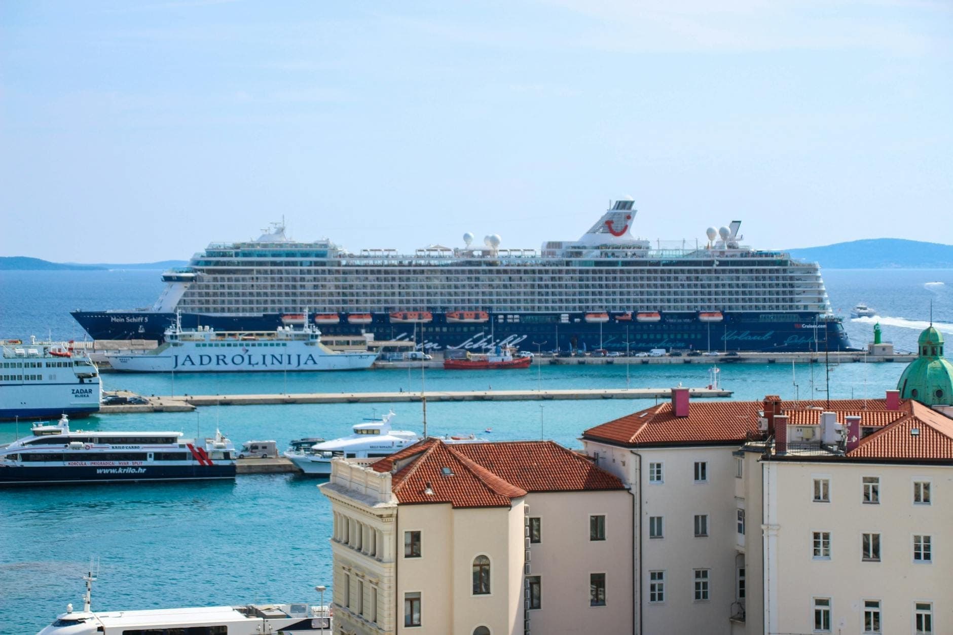 Vista panoramica del porto di Spalato con diversi traghetti, tra cui una nave di Jadrolinija, ormeggiati vicino al centro storico e navi da crociera sullo sfondo.