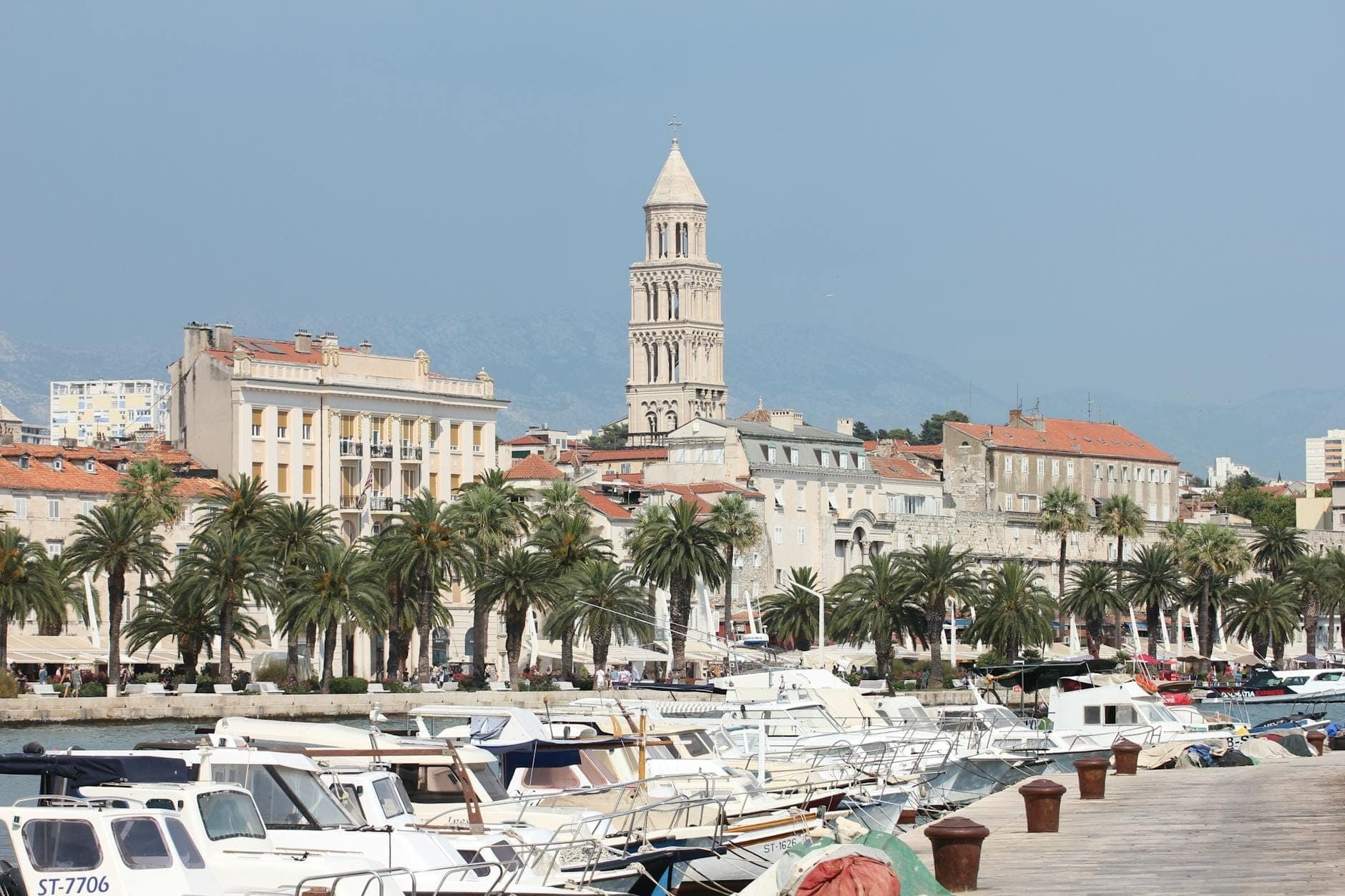 Lungomare di Spalato con un affollato terminal dei traghetti e barche ormeggiate davanti al viale alberato di palme, con il campanile del Palazzo di Diocleziano visibile sullo sfondo.