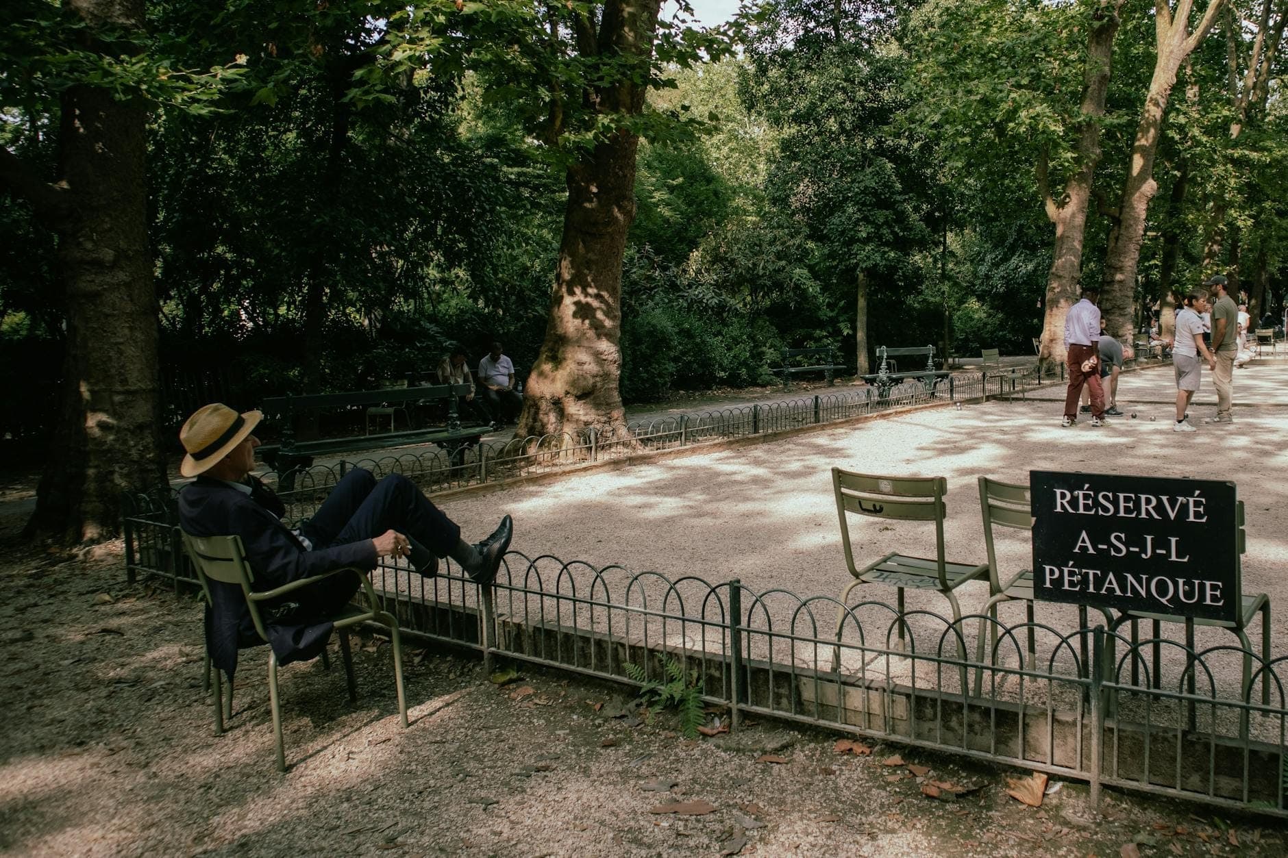 Shaded pétanque court in a leafy park with people relaxing and playing, a person in a hat lounging on a chair and a sign marked 'Réservé pétanque'.