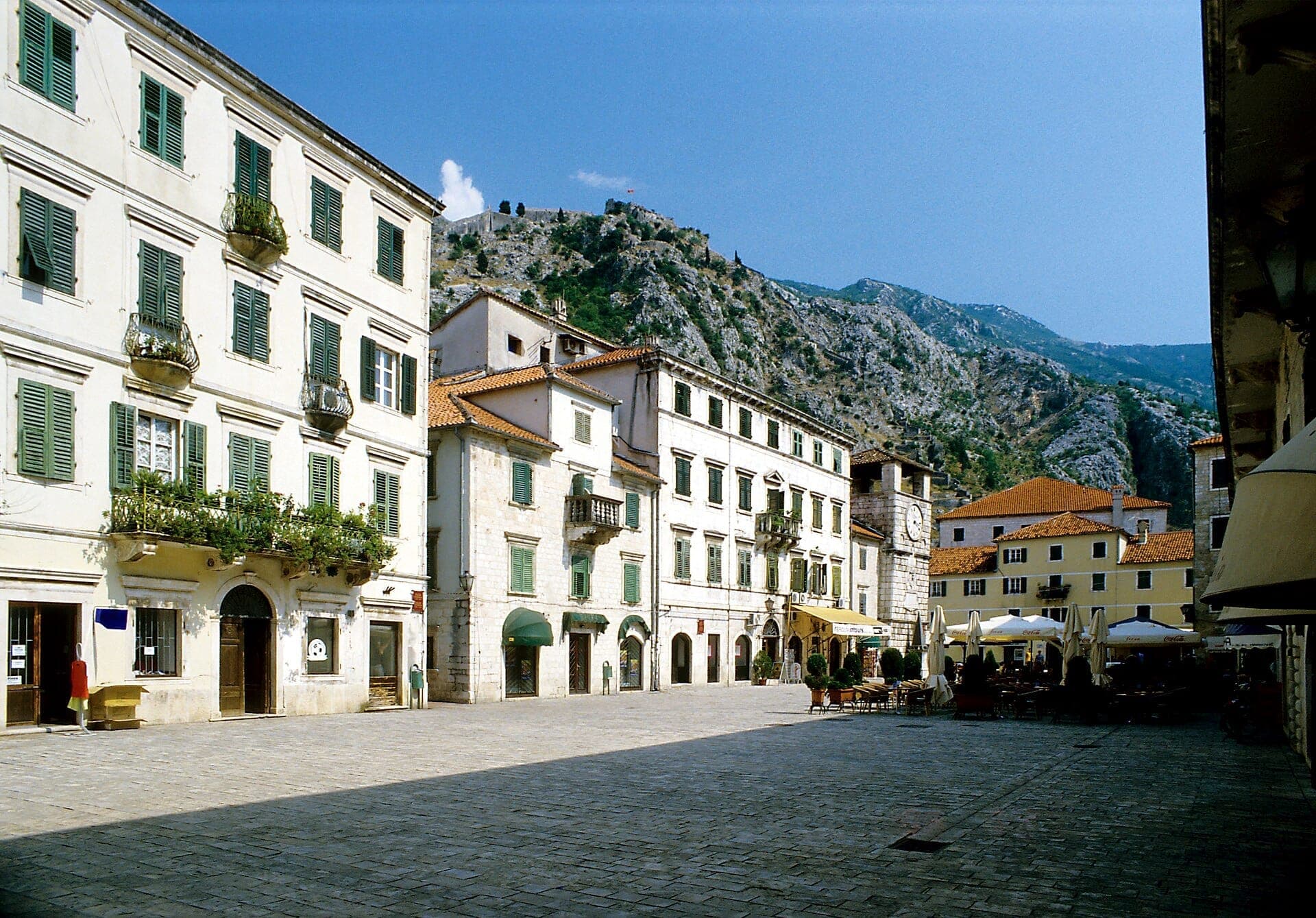 Vue panoramique de la Place d'Armes à Kotor, entourée de bâtiments historiques en pierre avec des volets verts et des montagnes en arrière-plan.