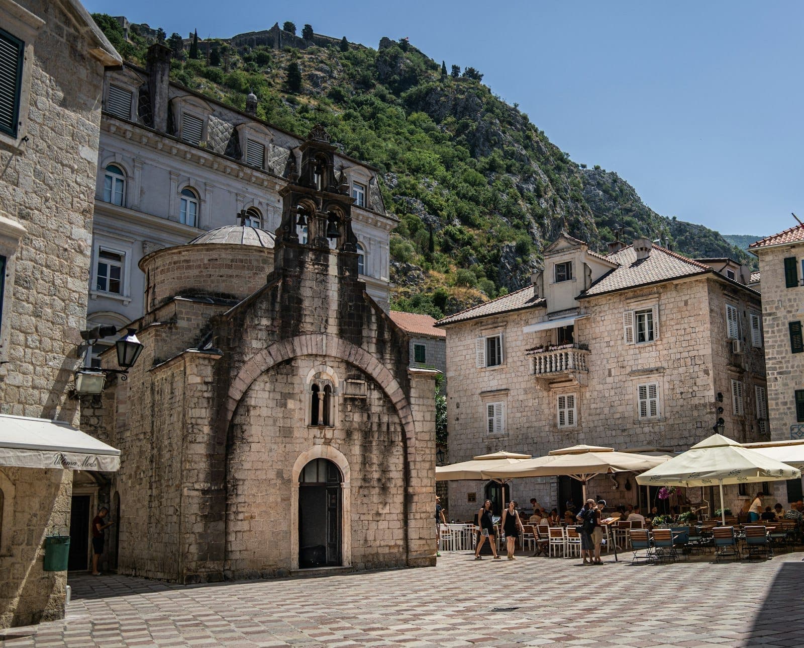 L'église Saint-Luc dans la vieille ville de Kotor, entourée de bâtiments en pierre, de cafés en plein air et de collines verdoyantes sous un ciel bleu dégagé.