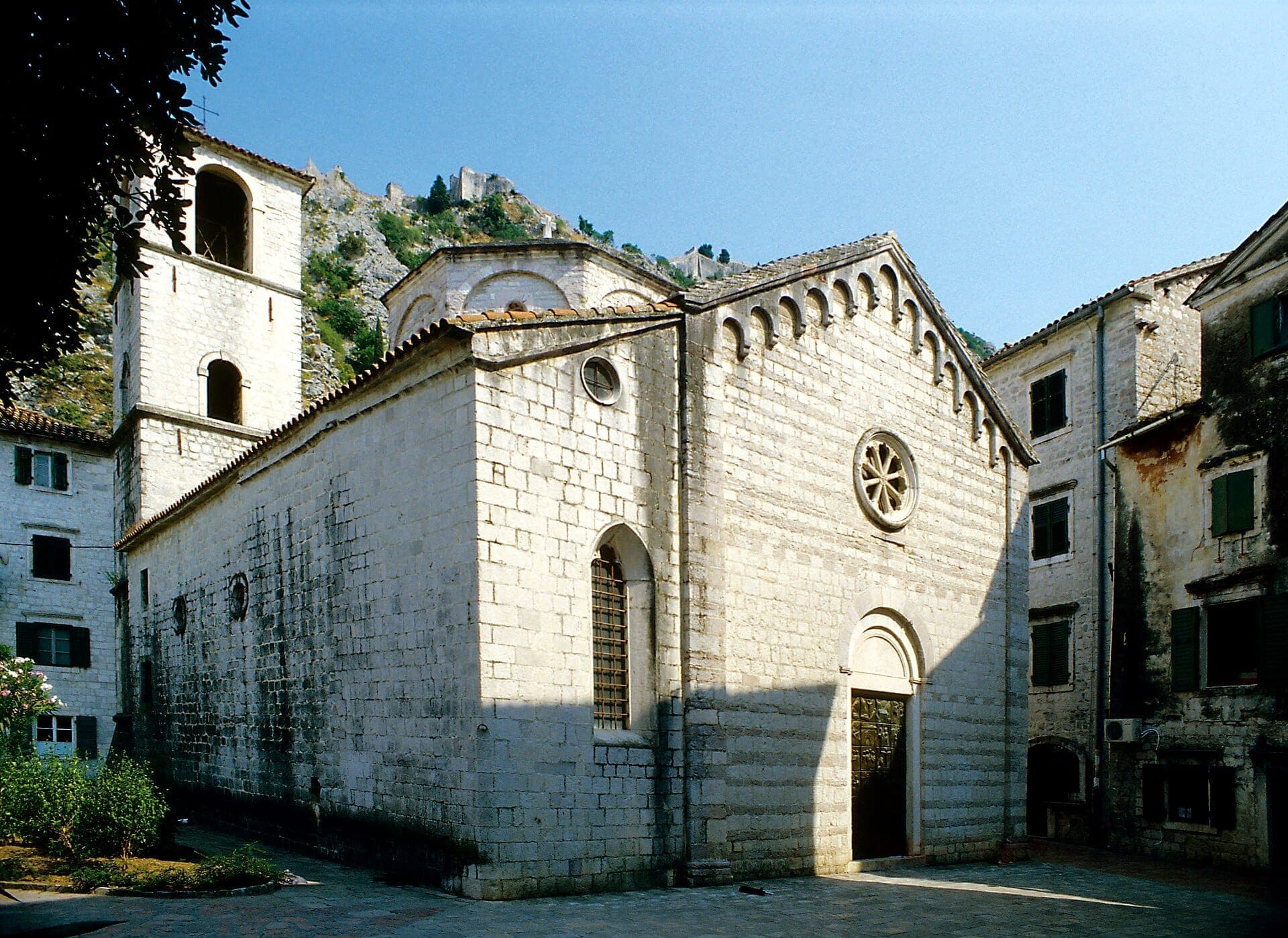 Vue extérieure de l'église collégiale Sainte-Marie à Kotor, mettant en valeur l'architecture romane en pierre, la rosace et le clocher sous un ciel bleu dégagé.