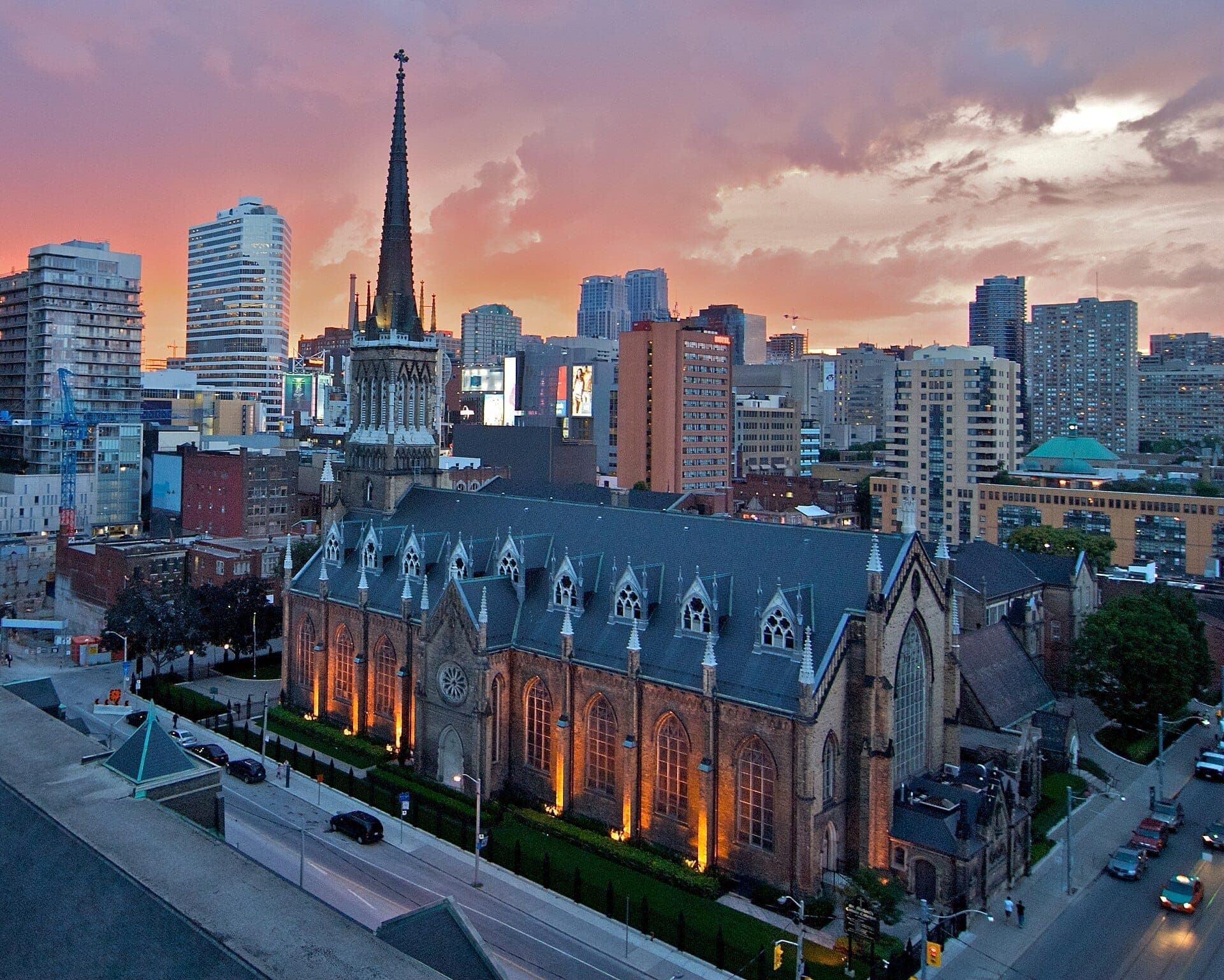 La Basilica Cattedrale di St. Michael's con la sua facciata gotica illuminata, l'alto campanile e le grandi finestre ad arco sullo sfondo dei grattacieli del centro di Toronto al tramonto.