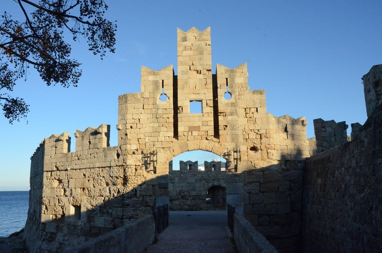 La Porte Saint-Paul à Rhodes, une porte médiévale en pierre avec des créneaux et des détails sculptés, se dresse sous un ciel bleu clair au bord de la mer.