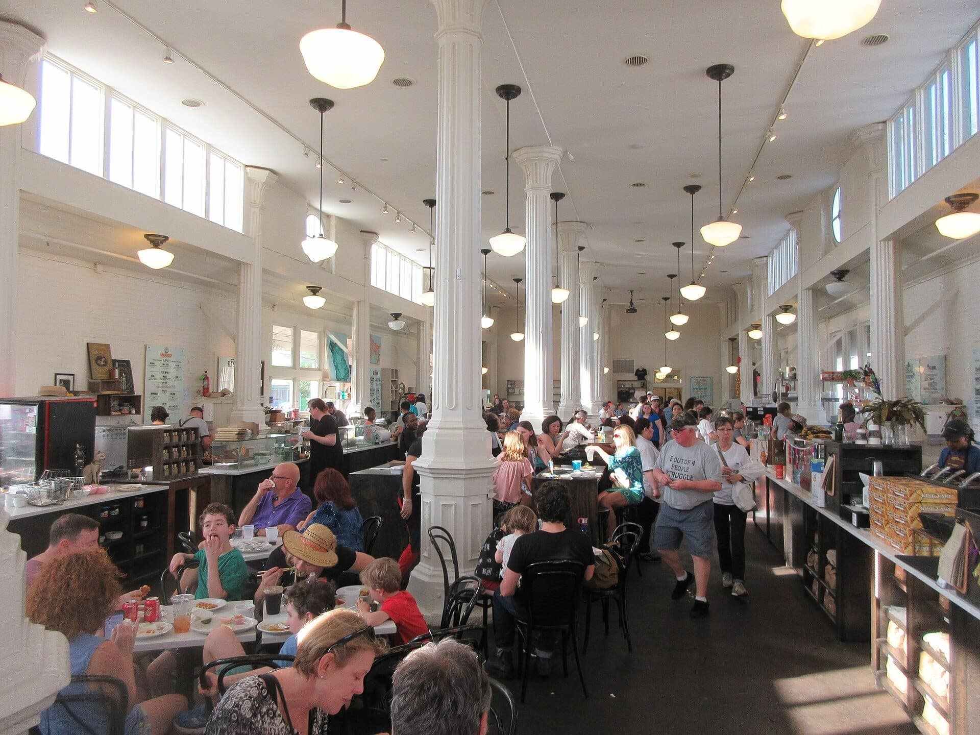 Interior movimentado do St. Roch Market com luz natural, altas colunas brancas, clientes em mesas comunitárias e vendedores atrás dos balcões em Nova Orleans.