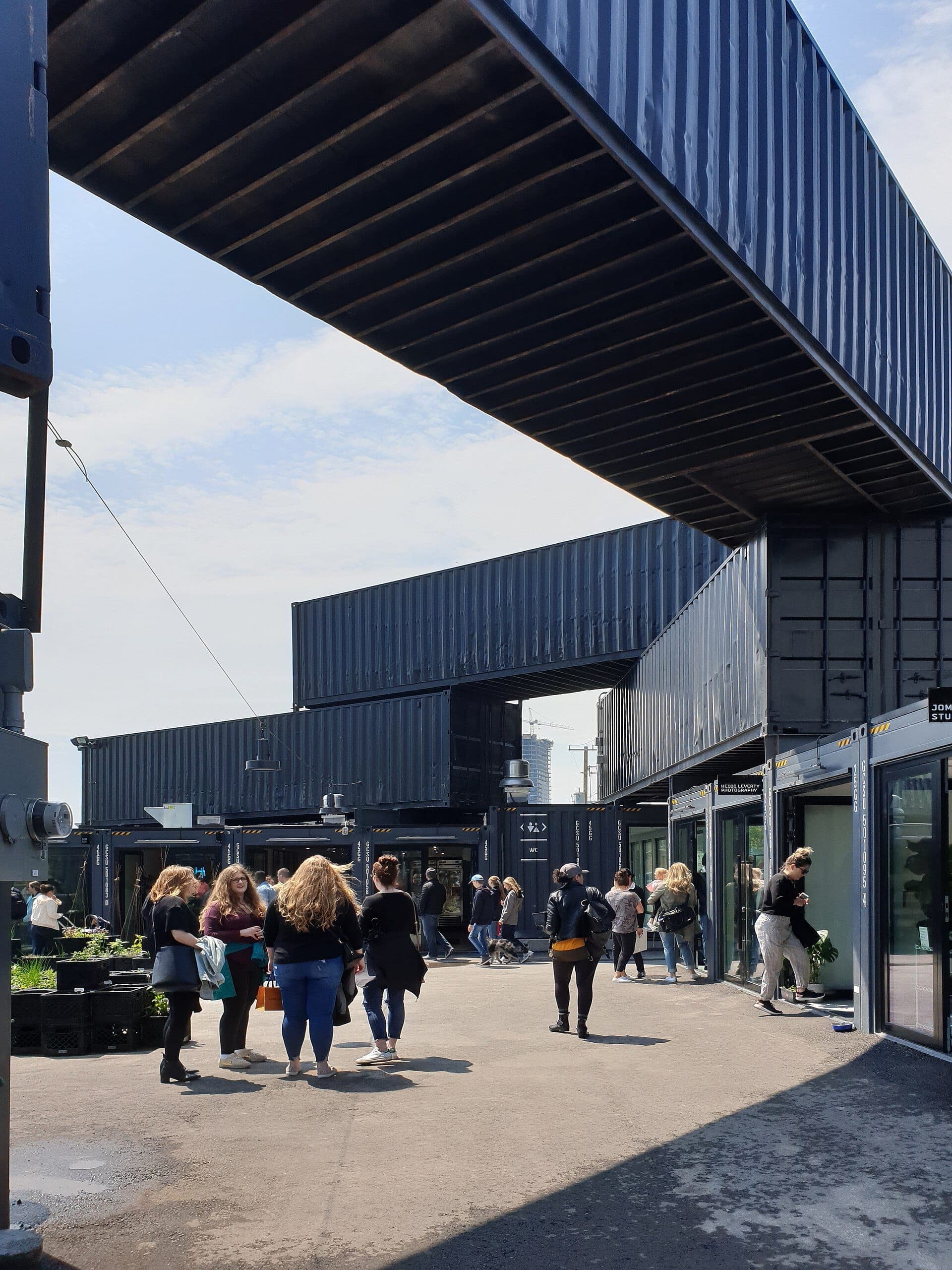 I visitatori camminano tra strutture di container neri sotto un cielo blu allo STACKT Market di Toronto, con negozi e porte in vetro visibili.
