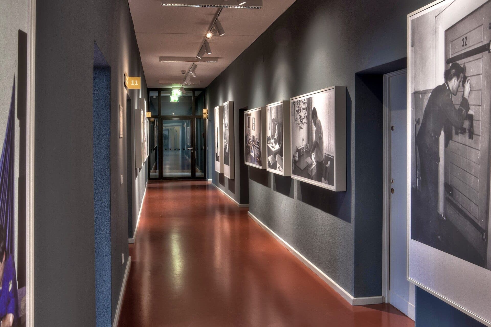 ทางเดินที่สว่างไสวใน Stasi Museum Berlin พร้อมภาพถ่ายวินเทจขาวดำที่จัดแสดงบนผนังสีฟ้าอมเทา