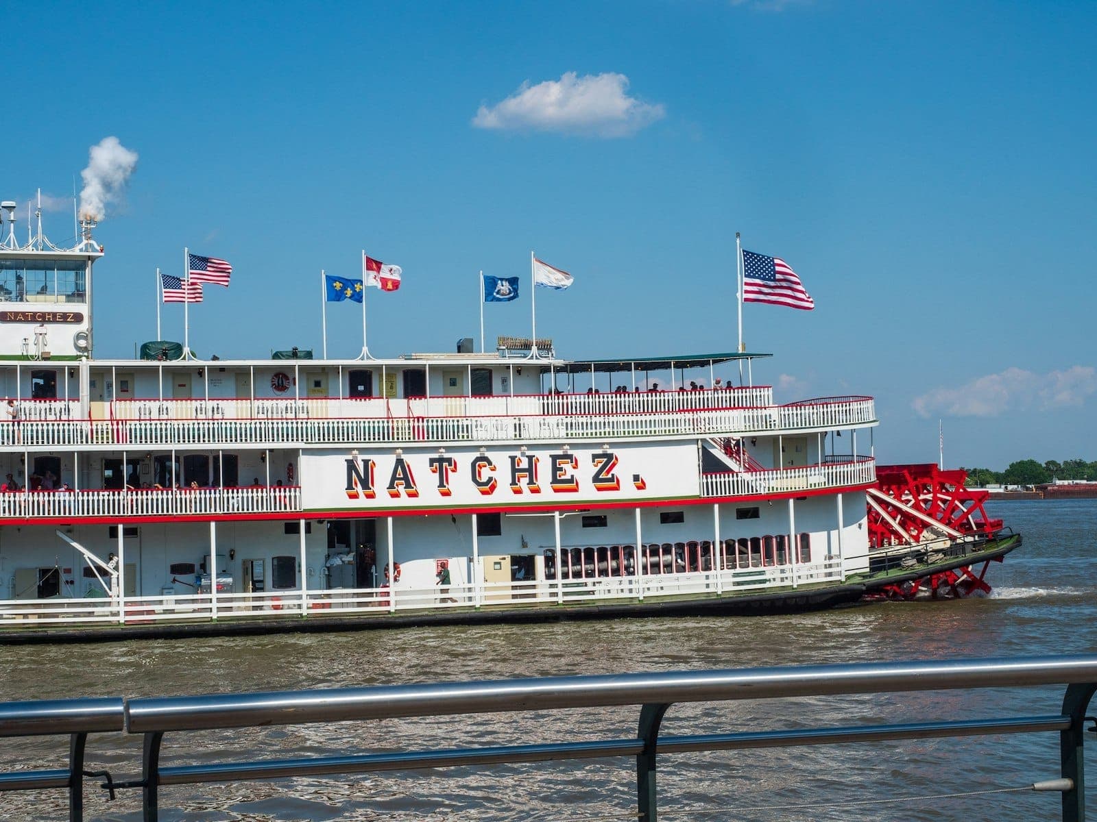 Steamboat Natchez cruising on the Mississippi River under a clear blue sky, with American flags and passengers visible on multiple decks.