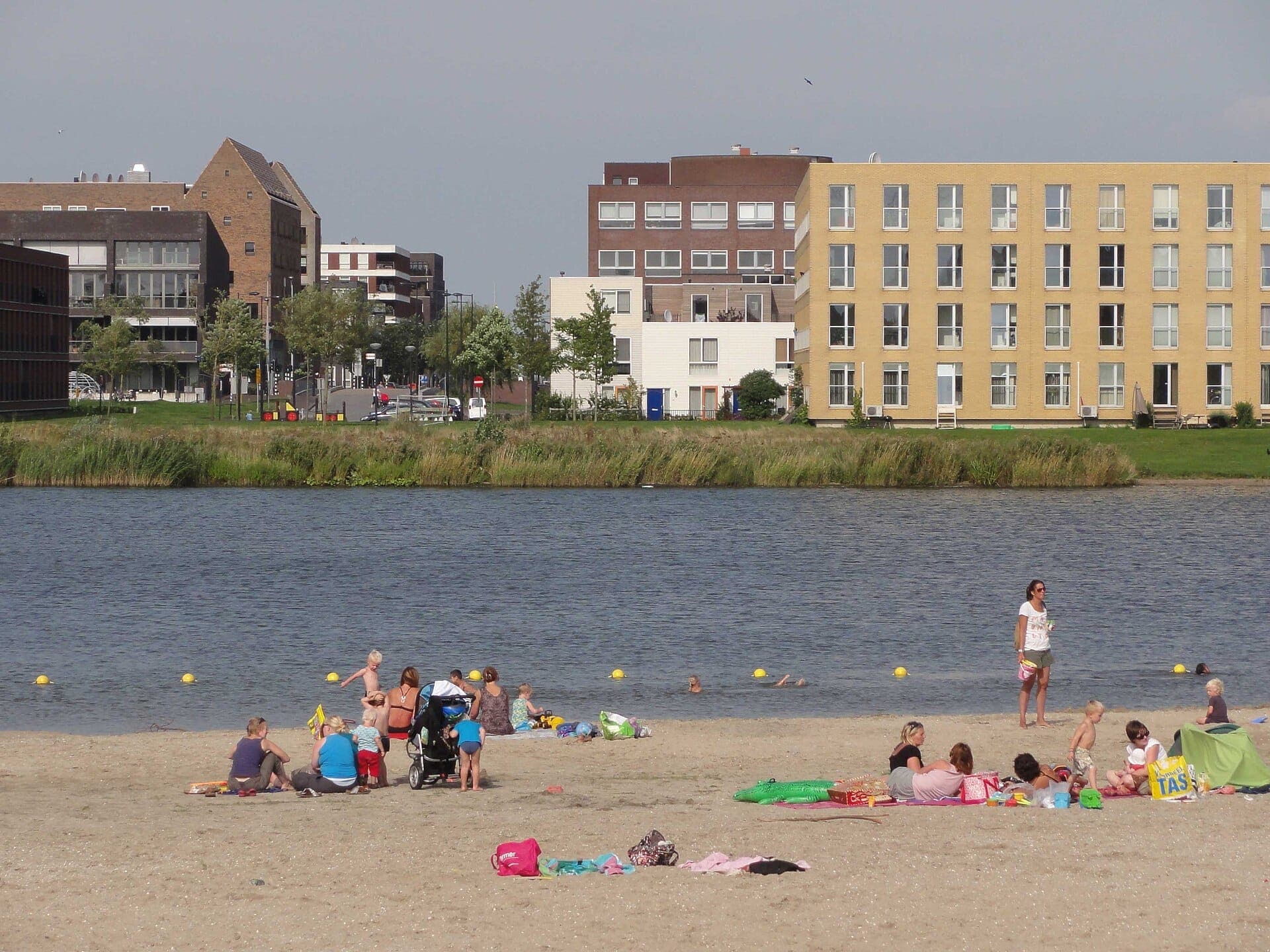 Familien und Kinder entspannen sich am Sandstrand von Strand IJburg, im Hintergrund moderne Apartmentgebäude und ruhiges Wasser.
