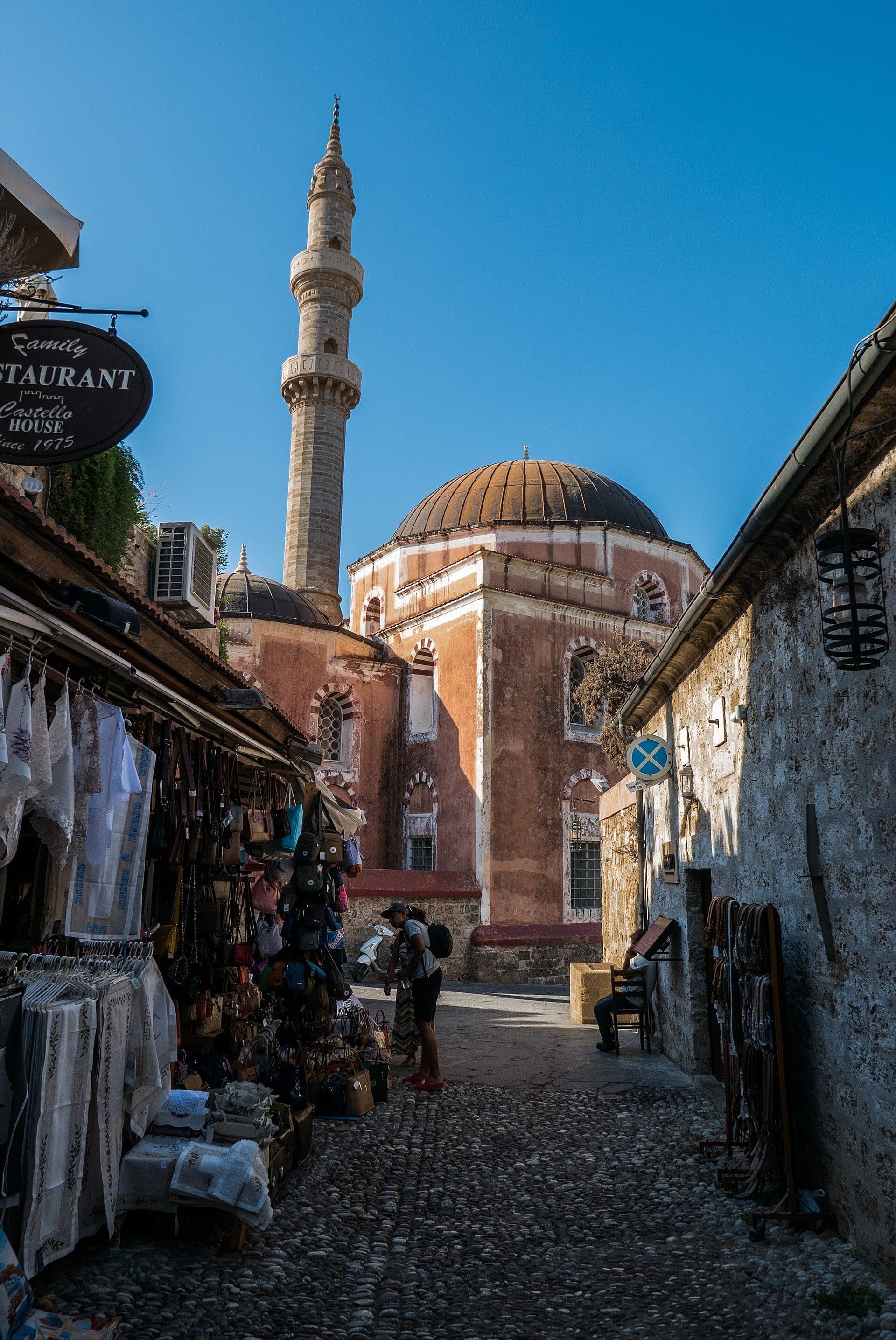La Mosquée de Soliman à Rhodes se dresse fièrement avec son dôme rose et son minaret, encadrée par les étals du marché de la vieille ville et un ciel bleu.