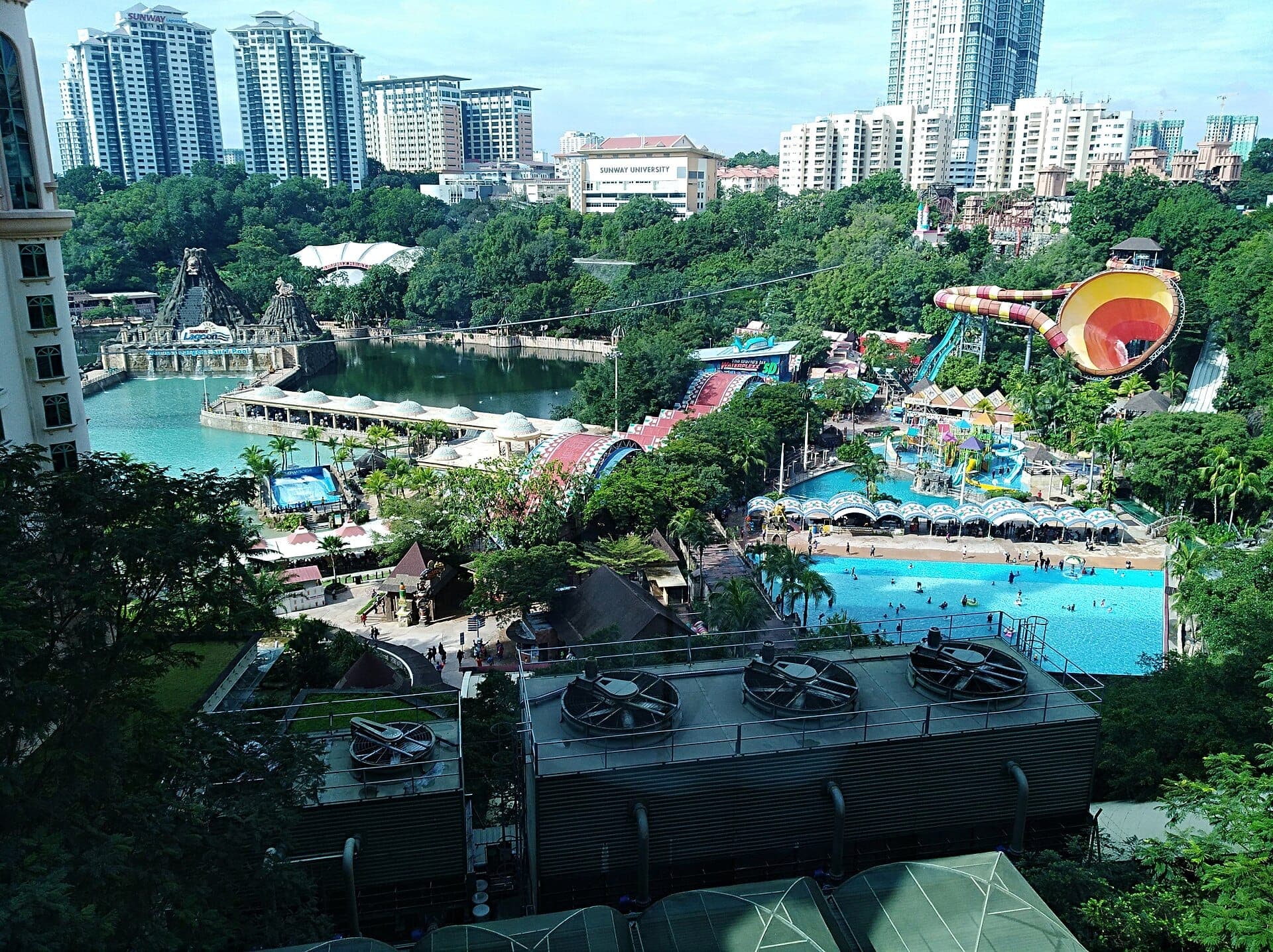 Kuala Lumpur'da havuzları, renkli su kaydırakları, yemyeşil bitki örtüsü ve çevresindeki yüksek katlı binalarıyla Sunway Lagoon tema parkının genel görünümü.