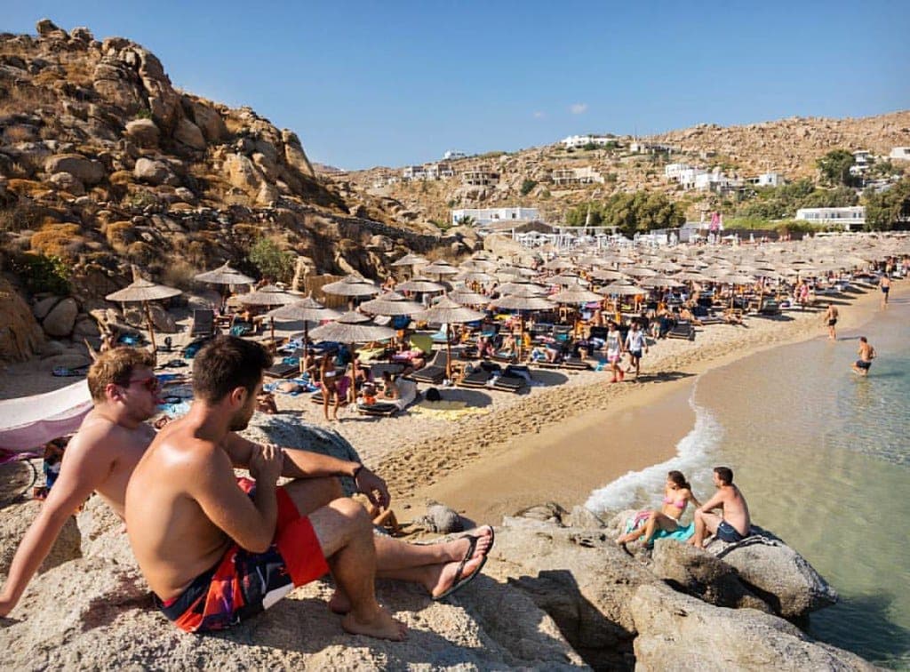 Blick auf den Super Paradise Beach auf Mykonos mit Liegestühlen, Strohschirmen, Menschen, die sich auf Felsen entspannen, und goldenen Hügeln unter einem klaren blauen Himmel.