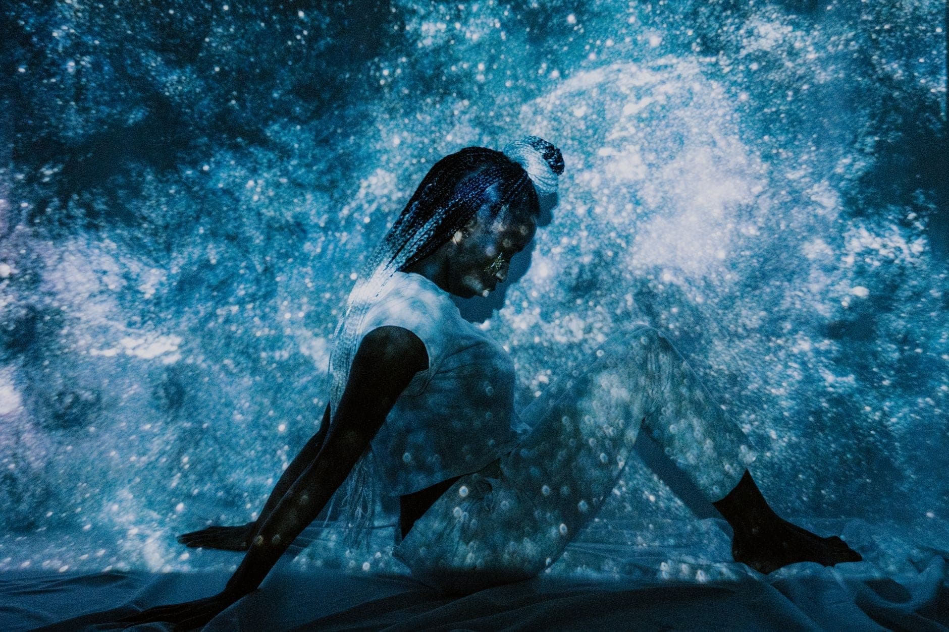Person in white sitting in a dark room surrounded by swirling blue and white light projections, creating a mesmerizing immersive art experience.