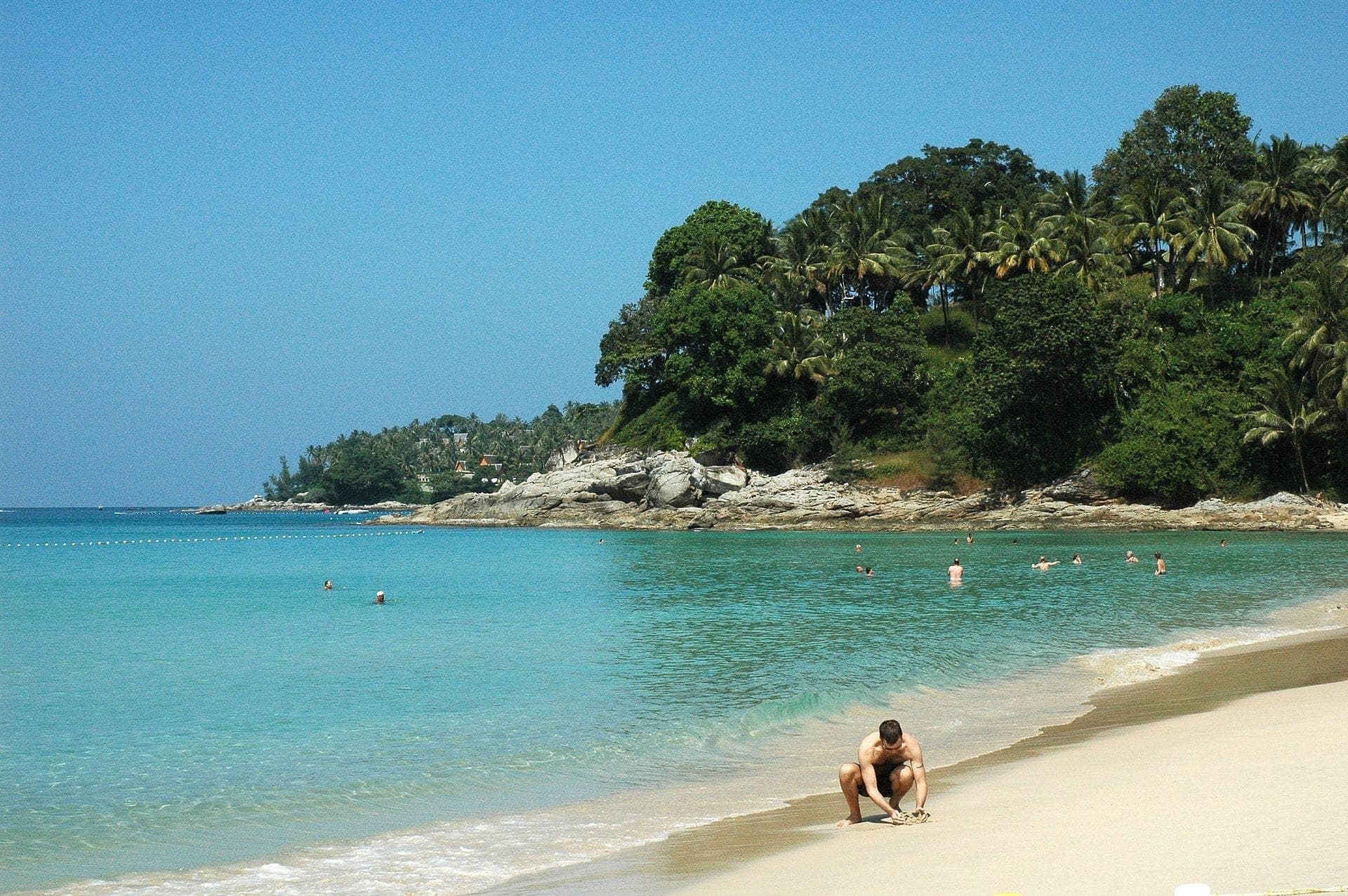 Surin Beach in Phuket with clear turquoise water, swimmers in the sea, a man on the sand, and lush green trees lining the rocky shore.