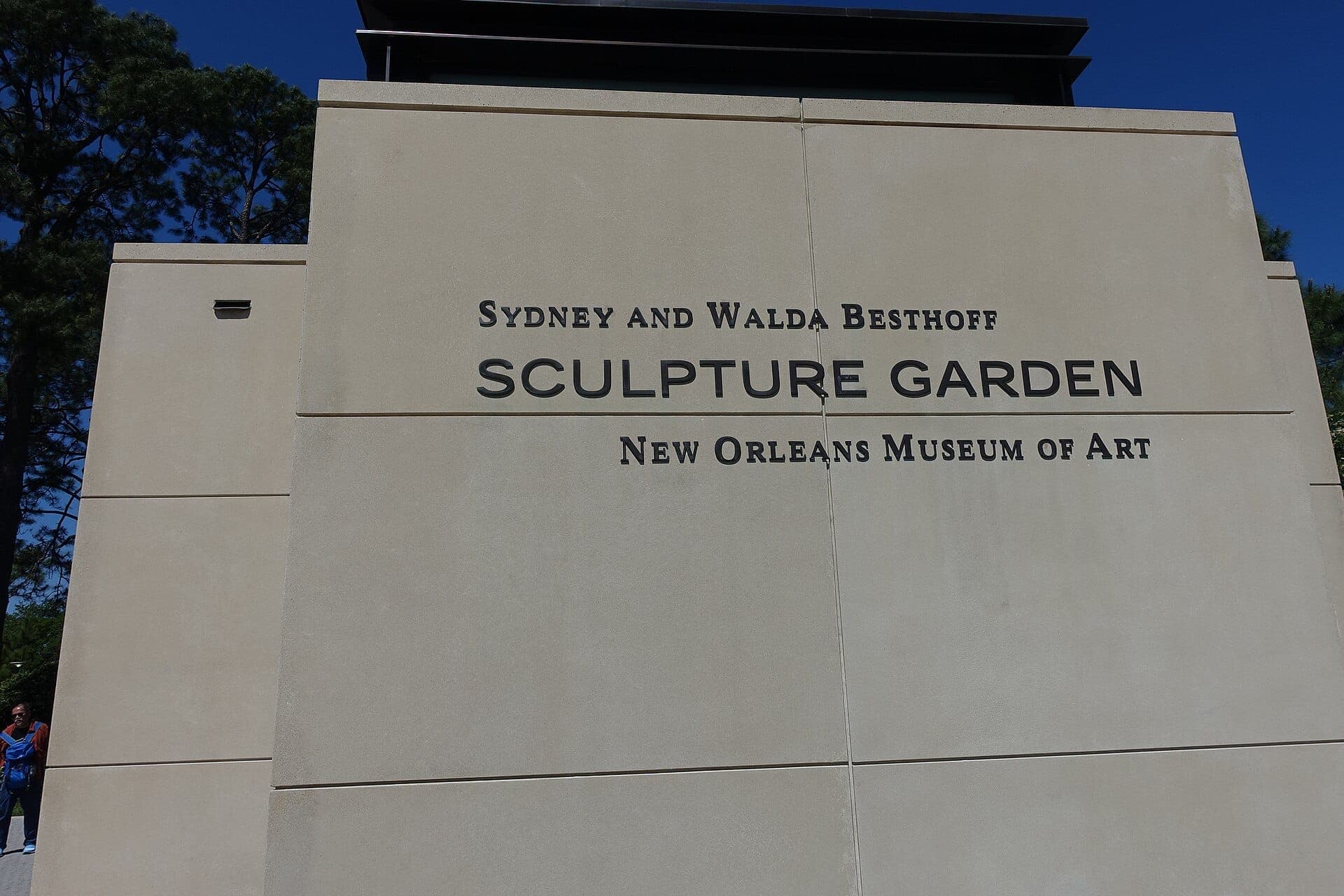 Entrance wall of the Sydney and Walda Besthoff Sculpture Garden with name signage, beige stone, and blue sky in the background.
