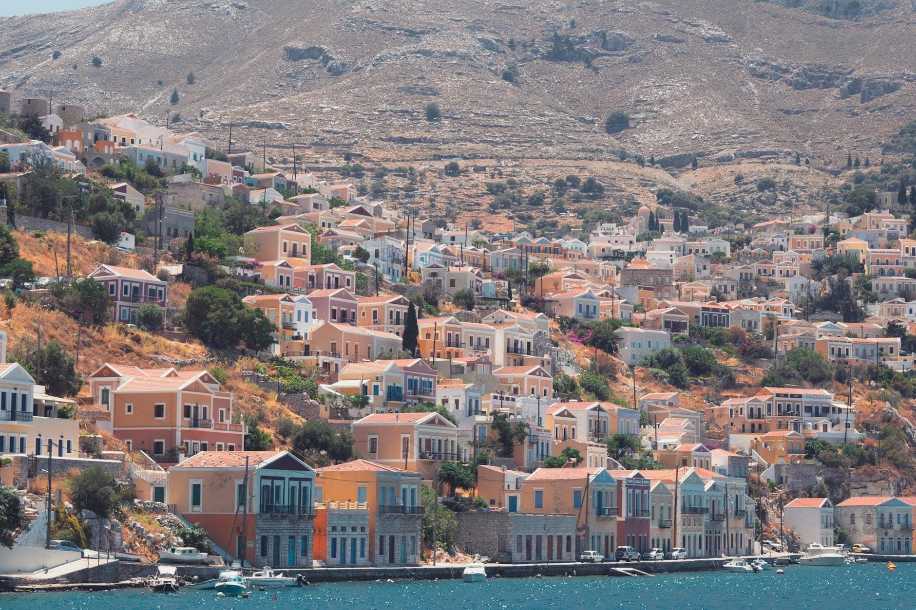 Demeures néoclassiques colorées étagées sur les collines de l'île de Symi, dominant un port d'un bleu étincelant avec en arrière-plan des montagnes arides.