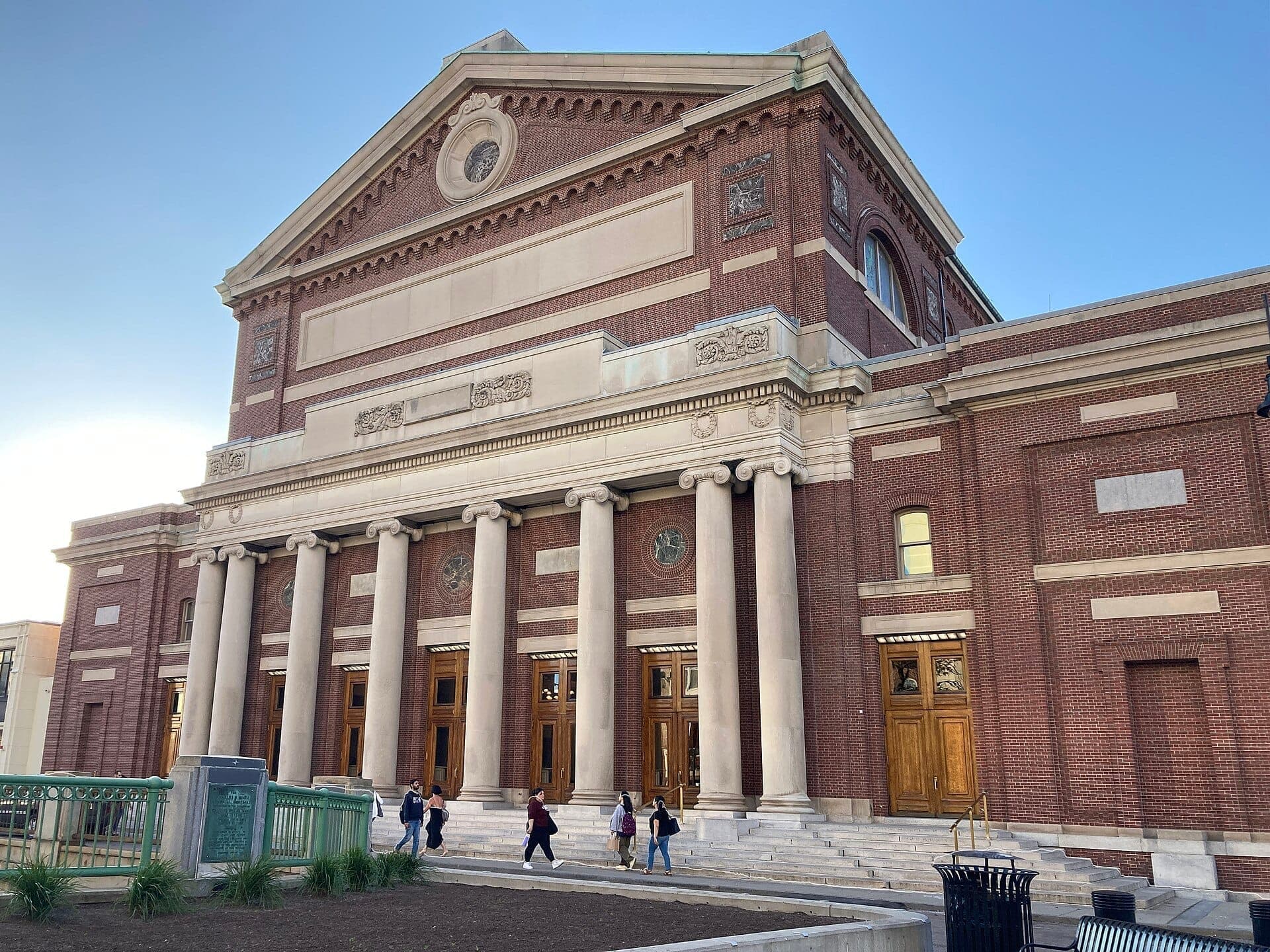 La façade néoclassique du Symphony Hall de Boston, avec de hautes colonnes, des murs en briques rouges et des visiteurs montant les marches sous un ciel dégagé.