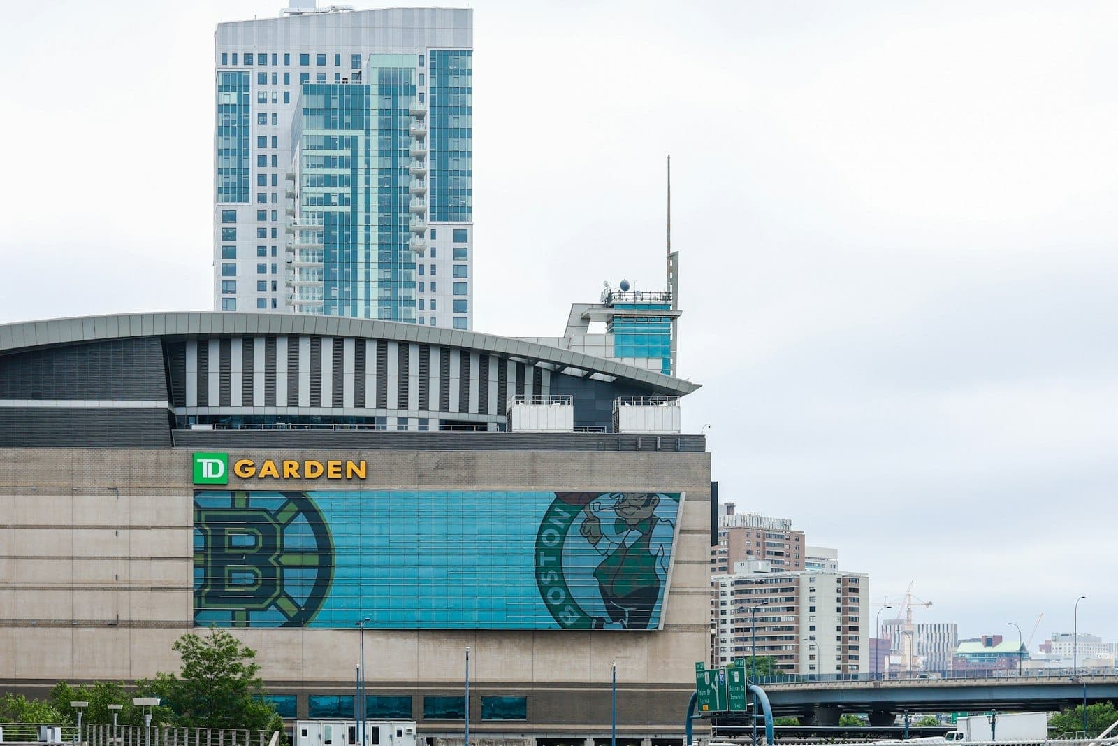 Vue de face de la salle TD Garden avec les logos des Celtics et des Bruins, sur fond d'immeubles modernes et d'un ciel nuageux de Boston.
