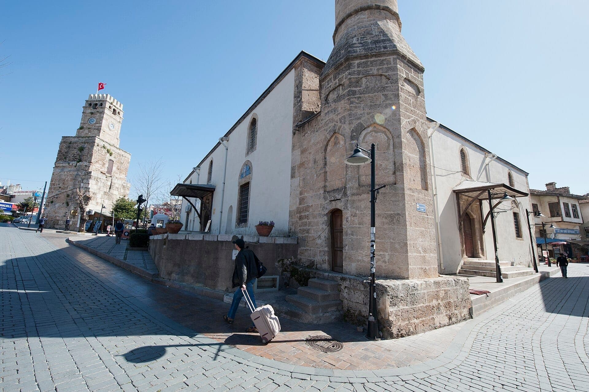 Die Tekeli Mehmet Paşa Moschee mit ihren Steinmauern und dem Minarett steht neben dem historischen Uhrturm von Antalya auf einer sonnigen Kopfsteinpflasterstraße.