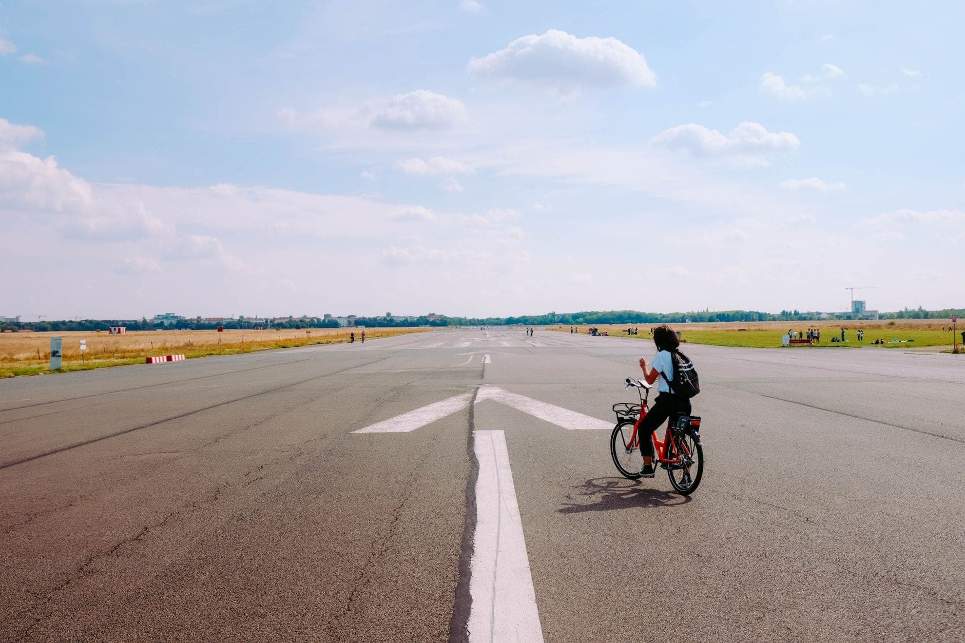 คนกำลังปั่นจักรยานบนรันเวย์กว้างโล่งกลาง Tempelhofer Feld ใต้ท้องฟ้าสีฟ้าพร้อมเมฆขาวประปราย
