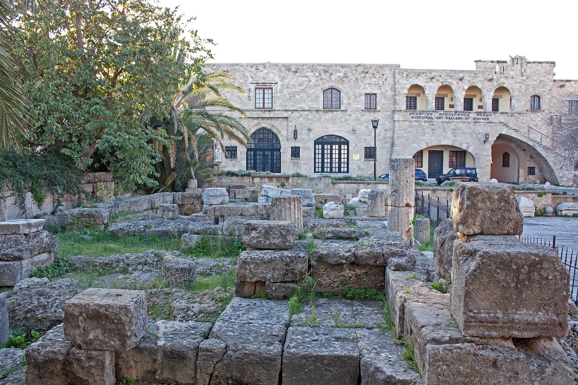 Ruines en pierre du Temple d'Aphrodite dans la vieille ville de Rhodes, avec des colonnes altérées et des blocs anciens entourés de verdure et de bâtiments historiques.
