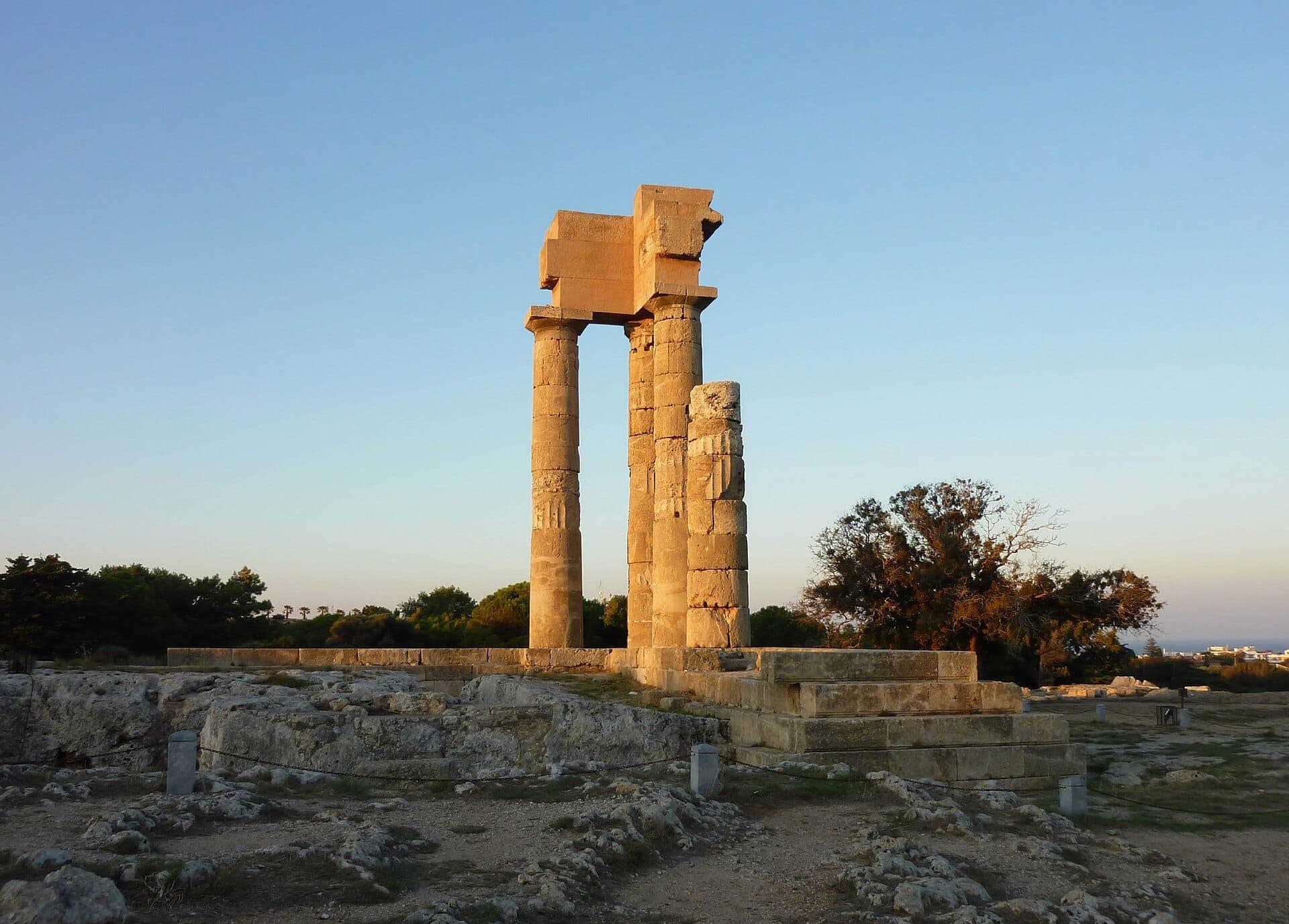 Ruines du Temple d'Apollon à Rhodes avec trois colonnes doriques debout, entourées de fondations en pierre et d'une végétation clairsemée sous un ciel bleu dégagé.