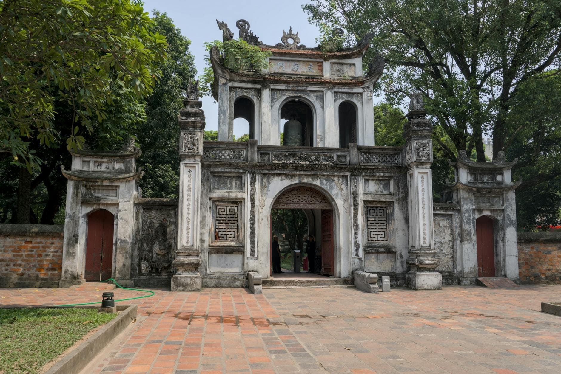 Szeroki widok na główną bramę Świątyni Literatury w Hanoi z tradycyjną wietnamską architekturą, otoczoną drzewami i ceglanym dziedzińcem w świetle dziennym.