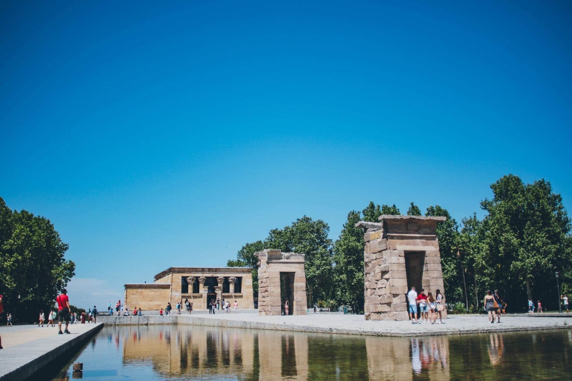 วิวกลางวันของ Templo de Debod ฟ้าสดใส วิหารหินสะท้อนบนผิวน้ำ นักท่องเที่ยวเดินชมรอบ ๆ และต้นไม้เขียวชอุ่มฉากหลัง