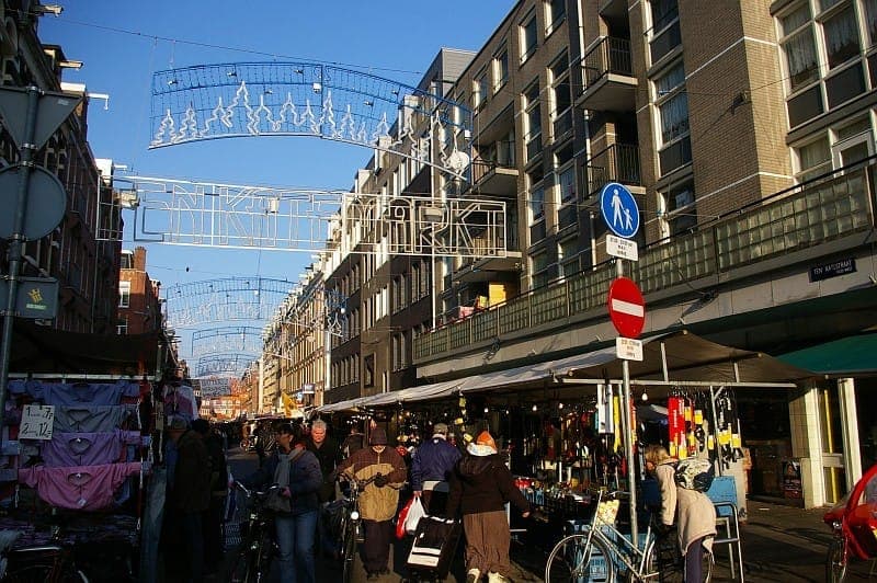 Einkäufer stöbern an Marktständen unter dem Ten Katemarkt-Schild in einer sonnigen Straße mit modernen Apartmentgebäuden in Amsterdam.