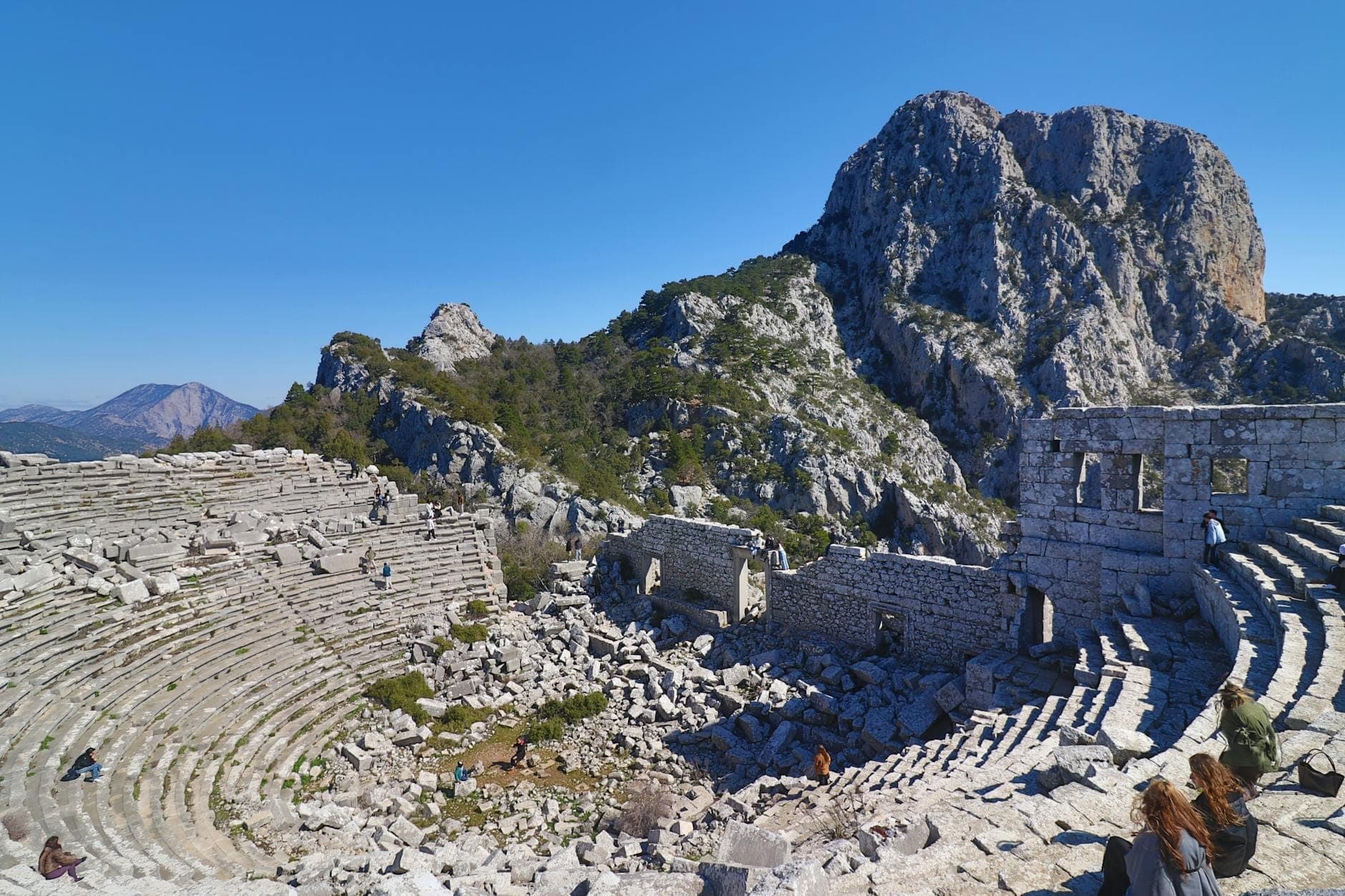 Das antike Steintheater von Termessos eingebettet in die dramatische Taurusgebirgslandschaft unter einem strahlend blauen Himmel, mit einigen Besuchern, die das Gelände erkunden.