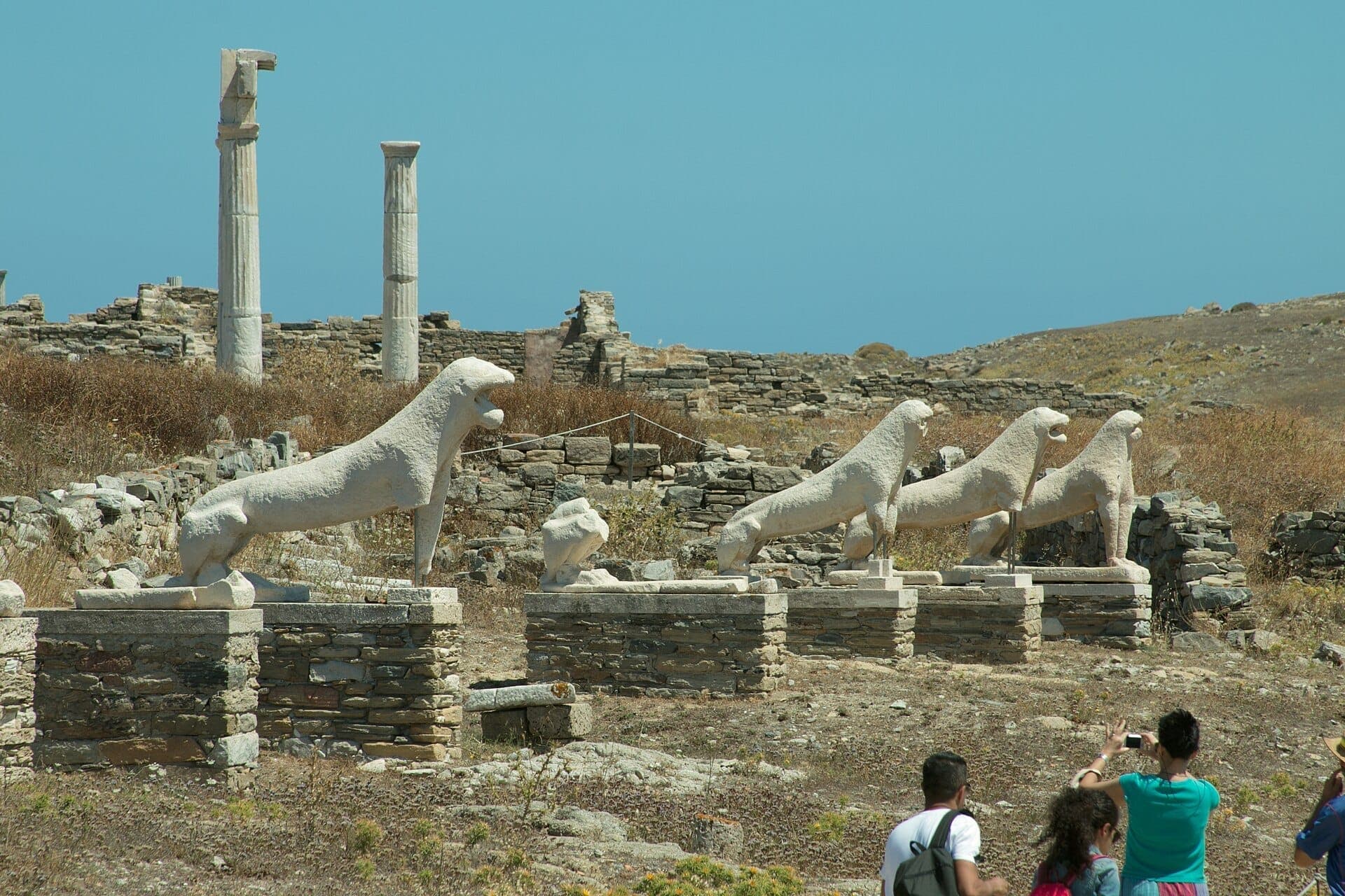 Reihe von Marmor-Löwenstatuen auf der Terrasse der Löwen, Delos, mit antiken Ruinen und Besuchern unter einem strahlend blauen Himmel.