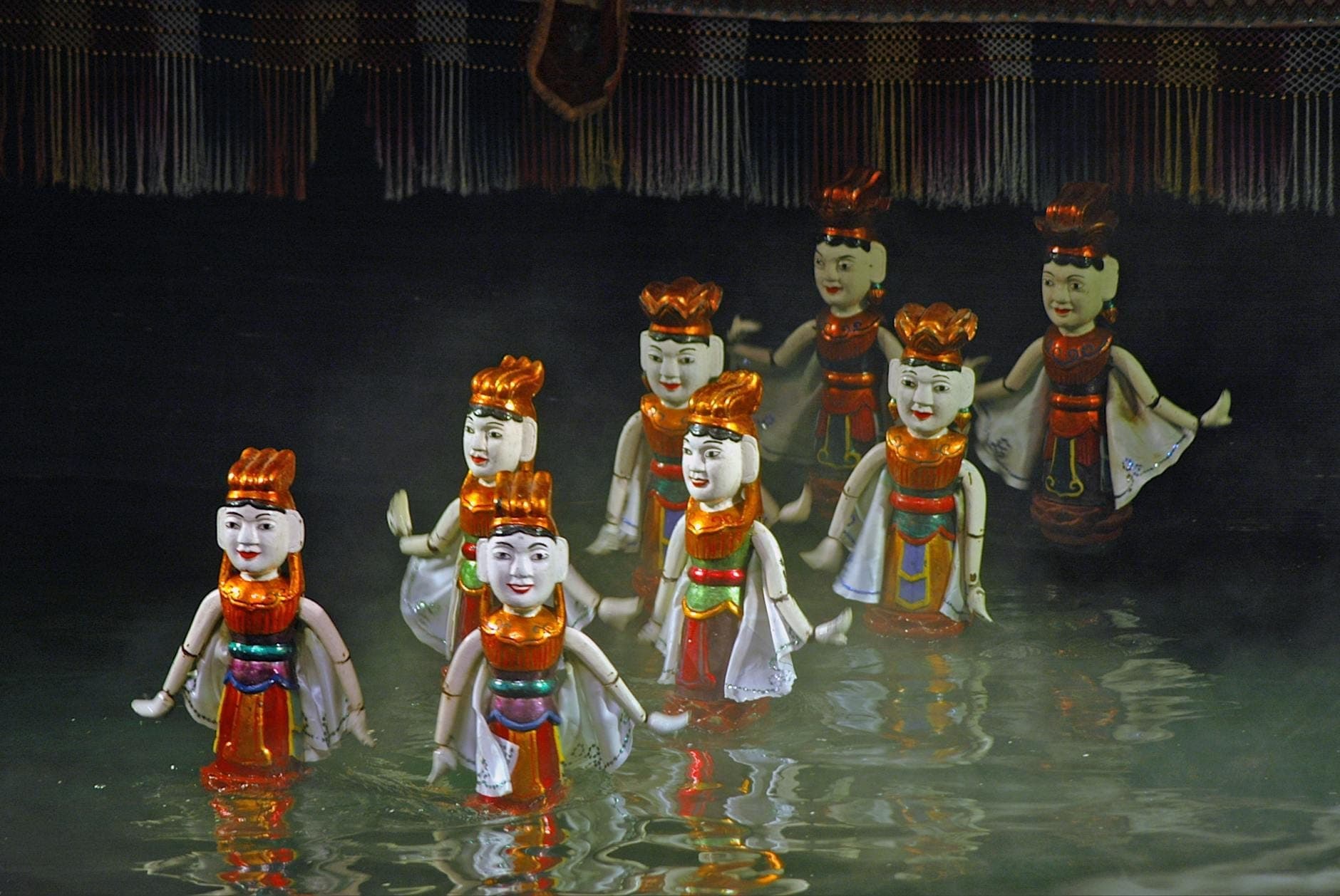 Grupa tradycyjnych wietnamskich lalek wodnych występująca na mglistej wodnej scenie z ciemnym tłem w Teatrze Lalek Wodnych Thang Long.