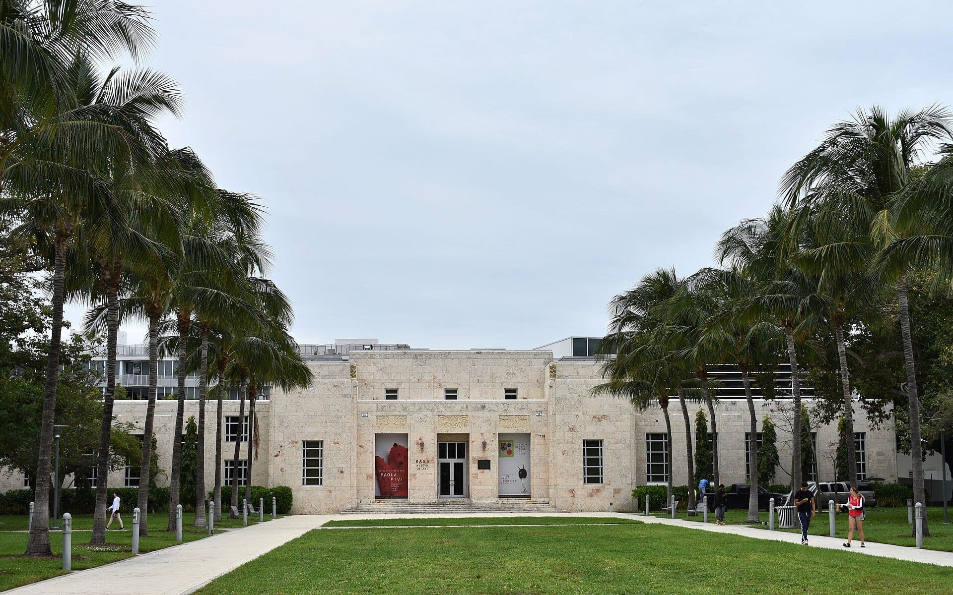 The Bass Museum in Miami Beach, a beige Art Deco building framed by tall palm trees and a wide lawn on a cloudy day.