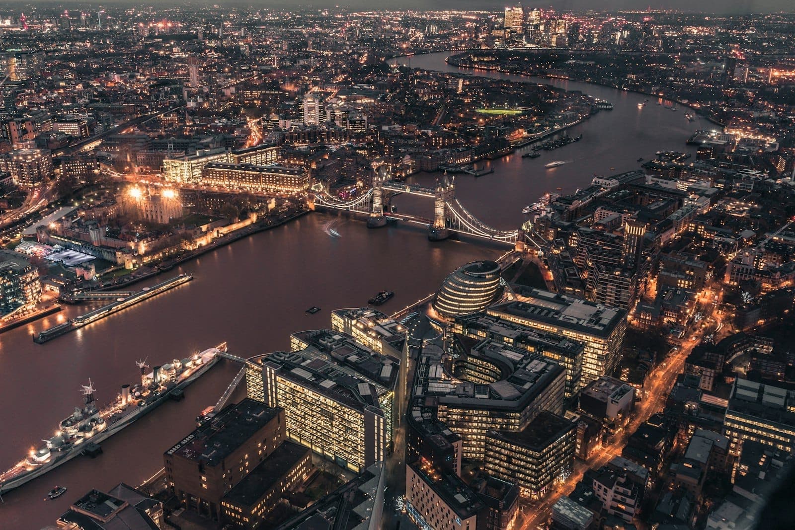 Cảnh nhìn từ trên cao vào ban đêm của The City of London với những tòa nhà văn phòng sáng đèn, cầu Tower Bridge và sông Thames uốn lượn qua kiến trúc hiện đại và lịch sử của Square Mile.