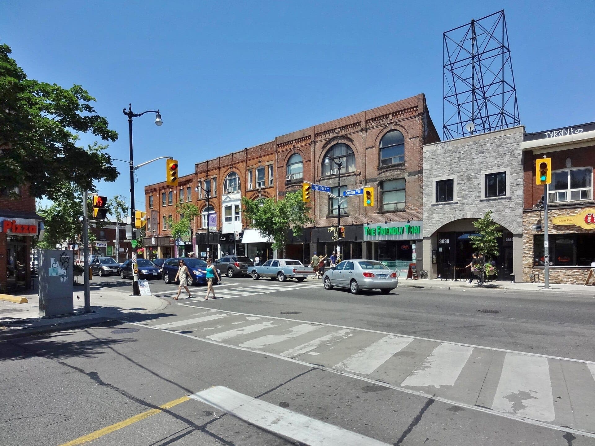 Una scena di strada soleggiata a The Junction, Toronto, con storici edifici in mattoni, auto e pedoni all'incrocio tra High Park e Dundas West.